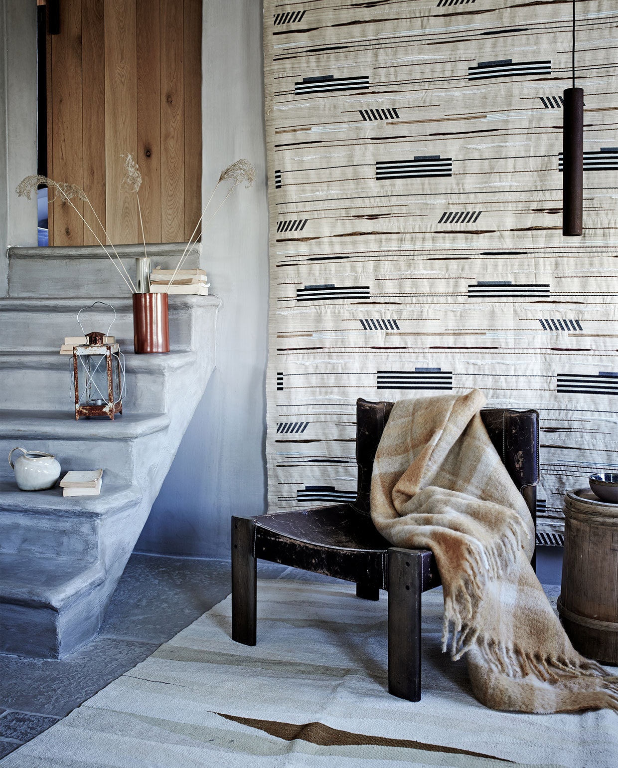 Stof wandkleed Pelops Gris Ombre van Nobilis, vloerkleed Vegetal Natural Stripes van Golran- (wva.nl). Vintage leren fauteuil- (pietjonker.nl). Mohair deken met ruit (zarahome.com). Kan, ecru vaas, schaaltje, alles van Serax (vtwonen.nl). Windlicht (com-anders.nl). Rode cilindervaas (klevering.nl). Houten bijzettafel (burbri.nl). Overig privébezit.