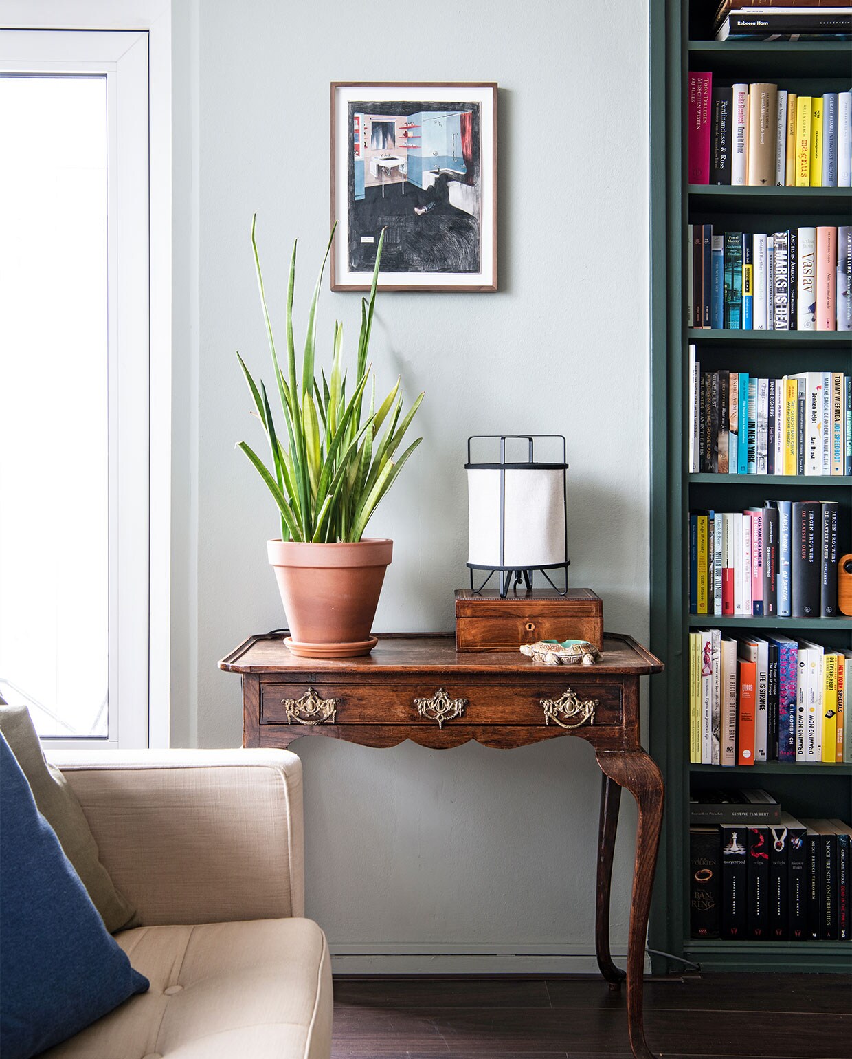 Vloer Ikea, muurverf Mizzle van Farrow &amp; Ball. Eettafel en oranje stoel cadeau gekregen, eettafelstoelen en hanglamp Gispen. Planttafel Marktplaats, vloerkleed Zara Home, televisiekast zelfgemaakt, fauteuil Rob Parry via Marktplaats. Kunst van links naar rechts Daniela Schwabe, Maze de Boer, Luis Xertu. Overig onbekend.