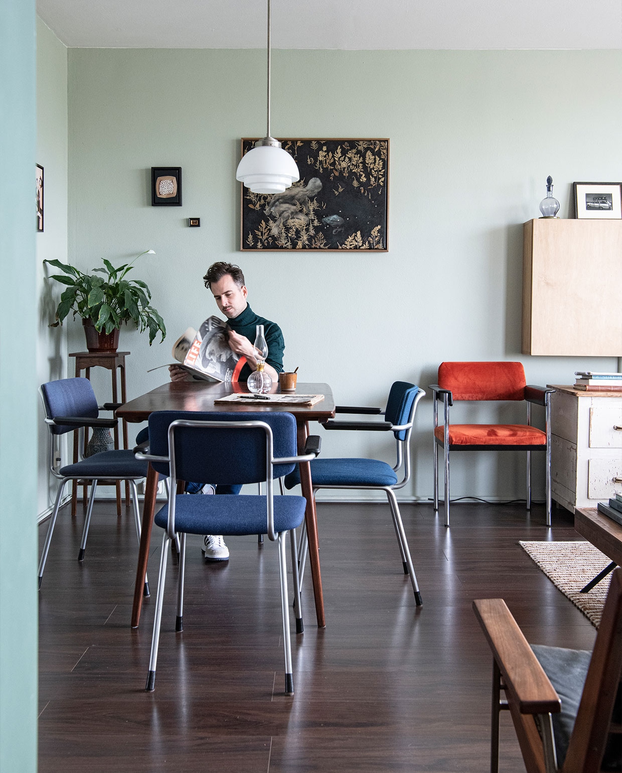 Vloer Ikea, muurverf Mizzle van Farrow &amp; Ball. Eettafel en oranje stoel cadeau gekregen, eettafelstoelen en hanglamp Gispen. Planttafel Marktplaats, vloerkleed Zara Home, televisiekast zelfgemaakt, fauteuil Rob Parry via Marktplaats. Kunst van links naar rechts Daniela Schwabe, Maze de Boer, Luis Xertu. Overig onbekend.