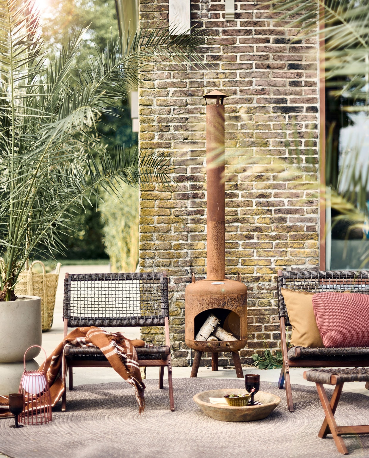 Klapstoelen Lois, per stuk € 149,-, voetstoel Lois € 75,-, beide van eucalyptushout met polyethyleen weefstructuur (basiclabel.nl). Buitenkleed, 140 cm (Ø) € 825,- (dacks.nl). Betonnen bloempot vanaf € 150,- (nordal.eu). Bruine wijnglazen, per stuk € 6,99 (zarahome.com). Tafellamp voor buiten van Lucide € 47,80 (vtwonen.be). Wollen plaid € 240,-, bruin buitenkussen € 25,- (paviljoennoord.com). Buitenhaard € 199,- (lanterfant.eu). Mosterdgele kom, 13 cm (Ø) € 12,-, gevlochten zeegras mand, 55×55 cm (hxØ) € 79,- (householdhardware.nl). Overig privébezit.