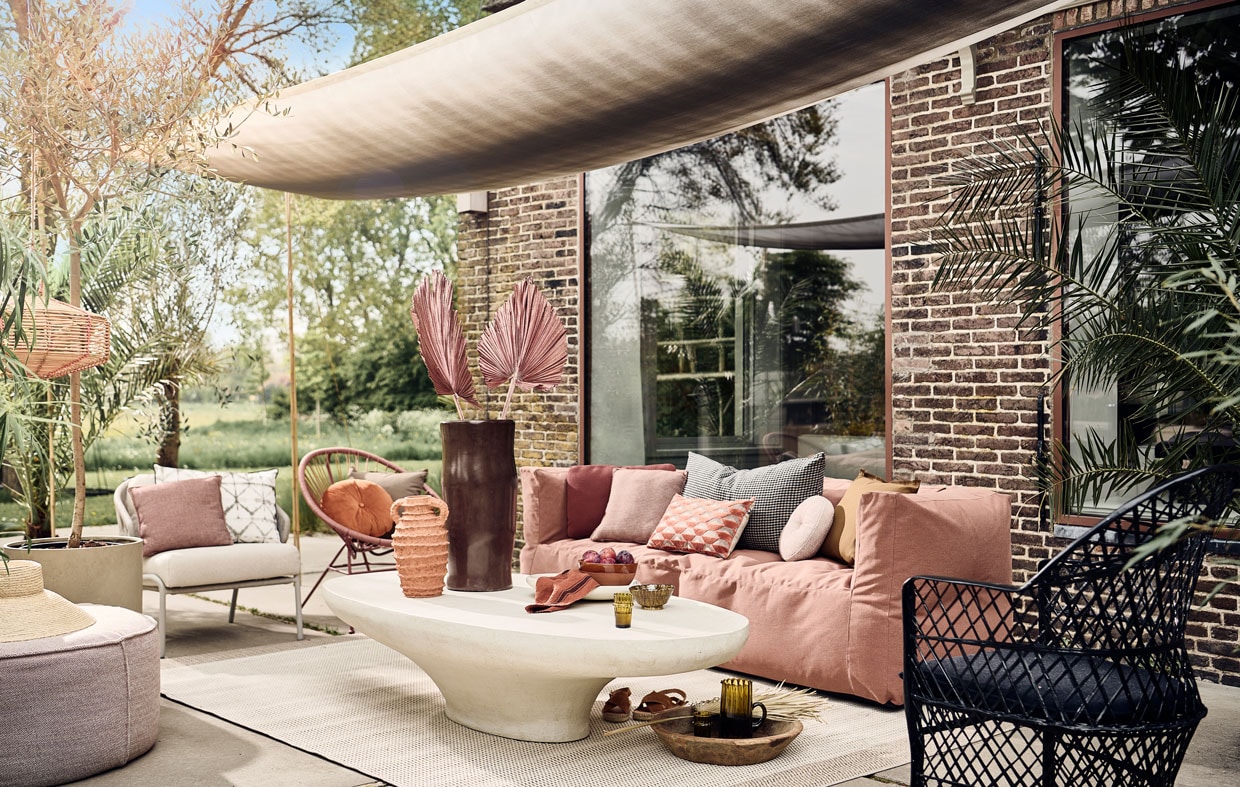 Schaduwdoek gemaakt van buitenstof Padua van Designers Guild, per meter € 73,-, wit rechthoekig buitenkussen met ikat print € 70,- (wva.nl). Buitenpoef vanaf € 525,-, zachtgrijze fauteuil met witte zitting € 2620,-, terra vierkante tuinkussens, per stuk € 151,-, terra langwerpig tuinkussen € 151,-, zachtroze rond tuinkussen vanaf € 410,- (dacks.nl). Hoed € 39,99 (zarahome.com). Rotan windlicht € 39,99, terracotta schaal, 20 cm (Ø) € 29,99, suède sandalen € 79,99 (sissy-boy.com). Betonnen bloempot vanaf € 150,- (nordal.eu). Buitenkleed van Lafuma, 200×290 cm € 269,90, oranje rond buitenkussen van Hartman € 24,95, buitenkussen met oranje retro dessin van Hartman € 29,95 (vtwonen.nl). Bruine tadelakt vaas € 115,- terracotta vaas, 35 cm (h) € 85,-, witte saladeschaal, terracotta met tadelakt finish, 38 cm (Ø) € 47,50, mosterdgele kom van klei, 13 cm (Ø) € 12,-, glas, per stuk € 6,50, karaf € 39,-, polyester zwarte fauteuil € 429,- (householdhardware.nl). Microcement salontafel vanaf € 1499,- (lanena-home.com). Driezits buitenbank van Subrella stof, 223x80x75 cm (lxbxh) € 1450,- (bryck.nl). Geruit zwart beige kussen € 90,-, honingbruin buitenkussen met rafelrand € 25,-, beige buitenkussen met rafelrand € 25,- terra linnen theedoek € 11,- (paviljoennoord.com). Roze loungestoel Samantha van Kave Home, polyester en staal, 84x72x75 cm (hxbxd) € 126,- (vtwonen.be).