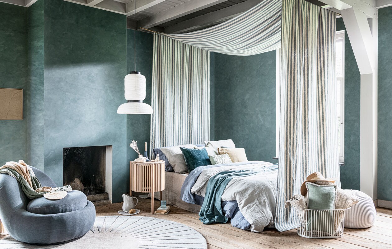 Kalkverf Blue Reef op de wand, per liter € 47,- (pure-original.nl). Schilderij met zandtextuur € 250,- (mooswillemsen.com). Fauteuil Babette, 84x93x73 cm (bxdxh) € 499,- (sofacompany.com). Wollen vloerkleed, 200 cm (Ø) € 399,- (made.com). Turkooizen-beige plaid € 139,-, poef € 430,- (homestede.nl). Bijzettafel 44×64 cm (Øxh) € 969,- (bolia.com). Hanglamp € 191,- (vantil.nl). Hemel gemaakt van stof Endless Summer van Barbara Osorio, 310 cm breed, per meter € 139,-, turkooizen velours kussen van William Yeoward € 98,-, turkooizen linnen plaid gemaakt van stof Canezza van Designers Guild, 297 cm breed, per meter € 94,-, opgerolde plaid van stof Theia van Nobilis, per meter € 238,- (wva.nl). Linnen laken in kleur Naturel, vanaf € 139,95, linnen dekbedovertrek in kleur Dusk Blue en Misty Green, vanaf € 199,95 (yumeko.com). Zachtgroen kussen met geborduurde bladeren € 22,99, zandkleurig kussen € 17,99, blauw linnen kussen € 19,99, zachtgroen kussen met witte bies € 22,99, gebroken wit kussen € 19,99, hoed € 22,99 (zarahome.com). Marmeren bord € 39,99, keramieken karaf € 29,99 (sissy-boy.com). Gebroken witte keramieken vaas € 36,- (serax.com). Houten kandelaar € 69,90 (letoile.store). Lichtblauw glas € 9,95 (anna-nina.nl). Metalen mand van Menu € 210,- (designtales.nl). Overig privébezit.