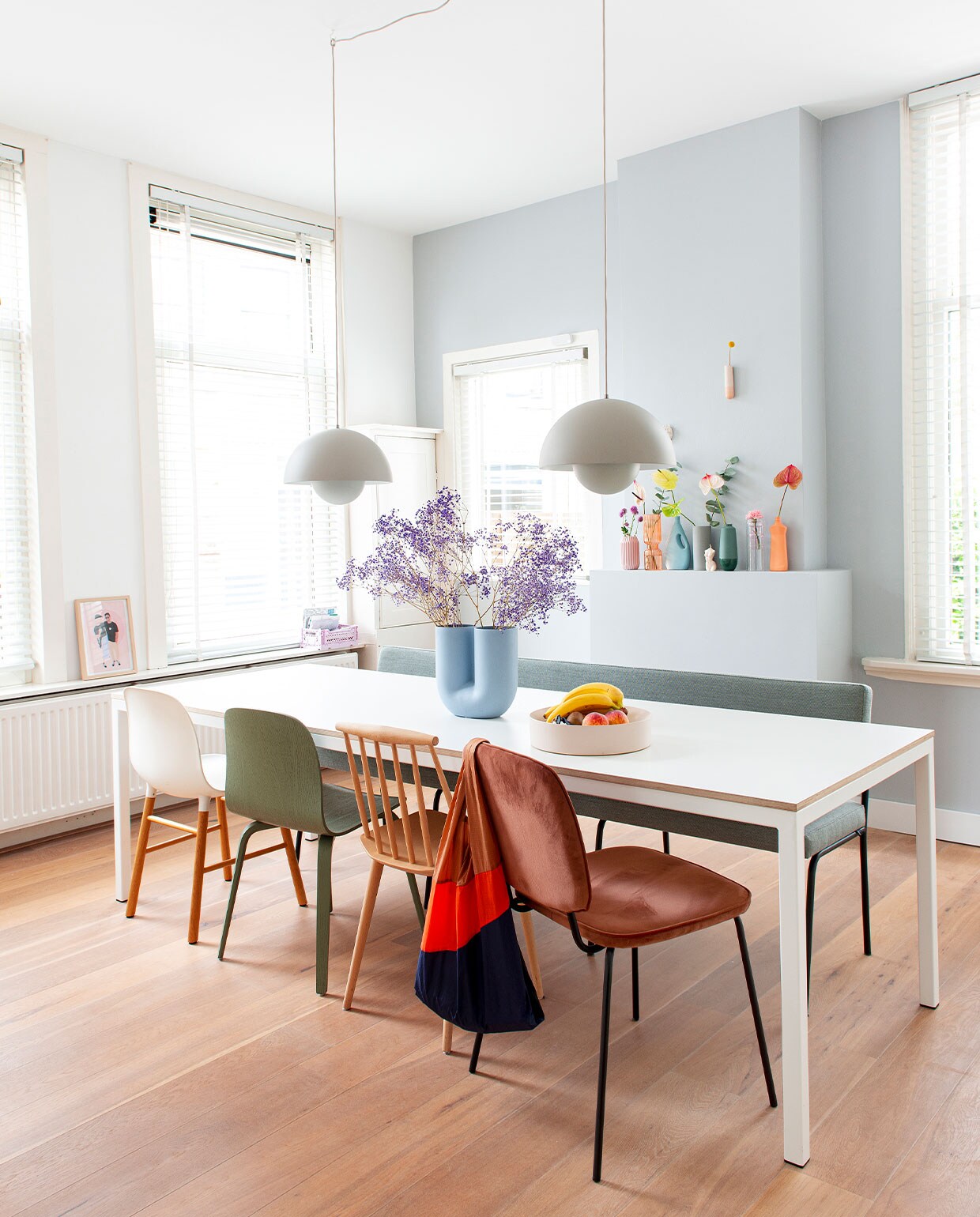 Muurverf Wild Dove van Flexa. Hanglampen FlowerPot van Verner Panton via &amp;Tradition. Tafel op maat gemaakt. Vaas Kink Muuto. Fruitschaal Fest Amsterdam. Witte stoel Normann Copenhagen. Groene stoel Muuto via Femkeido. Spijlenstoel Hay. Oranje stoel House Doctor. Bank Monday Fest Amsterdam via Wonenmetlef. Illustratie op vensterbank Melissa van Roon, MVR Design. Vazen op schoorsteen: roze en oranje Loods 5, blauwe en oranje fles Foekje Fleur, grijs Ferm Living, groen Hella Duijs. Wandvaas Studio Harm &amp; Elke.