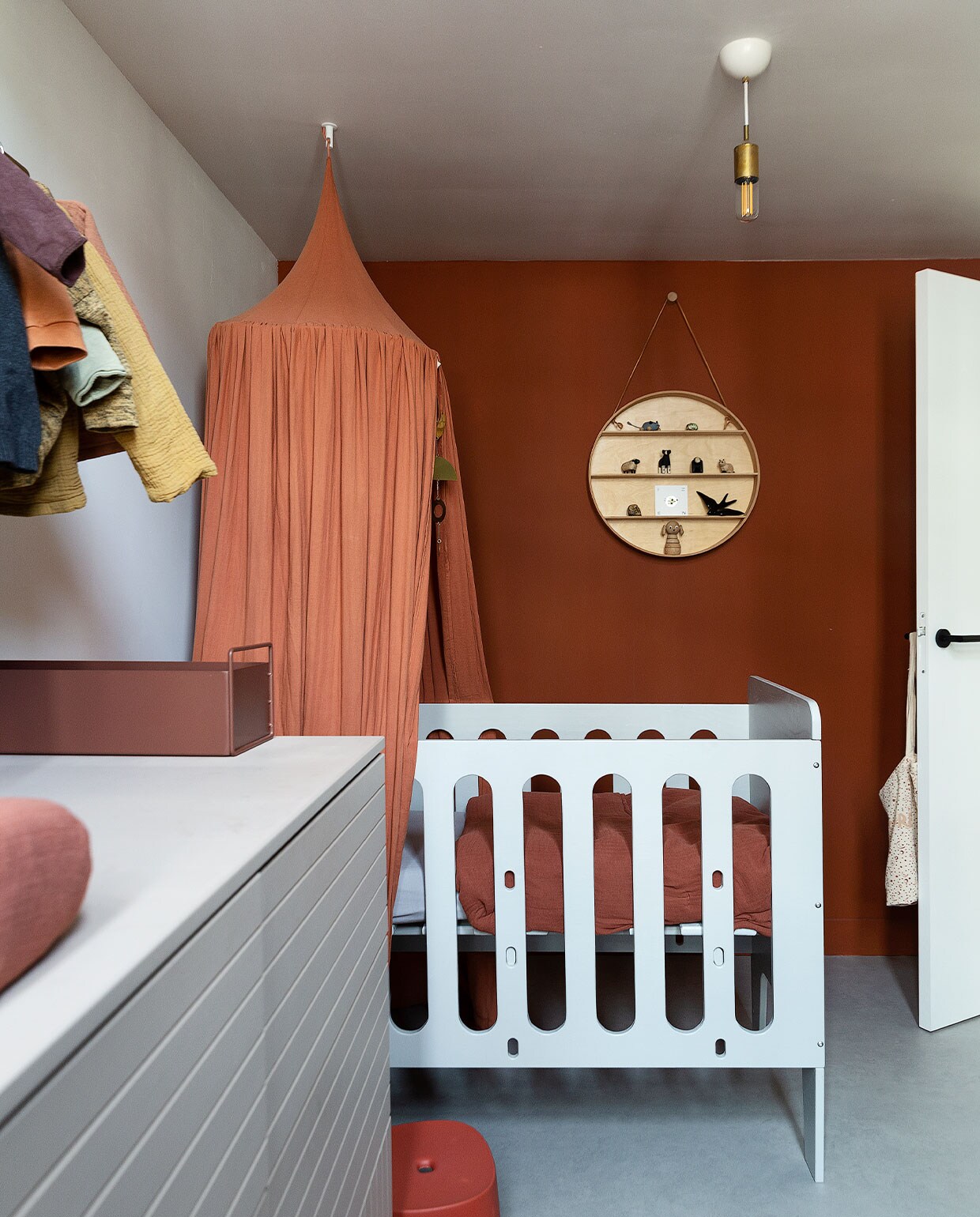 Babybed Timmer Toko van Overdijk, hemel Nobodinoz, beddengoed en aankleedkussen Garbo &amp; Friends. Rond wandobject met spullen van Mini &amp; Momo. Opstapkruk AliExpress, tas aan deurklink Archive Store. Commode eigen ontwerp op basis van keukenkasten Ikea, lijnen zelf erin gefreesd. Metalen bak Ferm Living. Plafondlamp Watt. Vloer Marmoleum van Forbo. Muurverf Pure Sienna van Flexa.