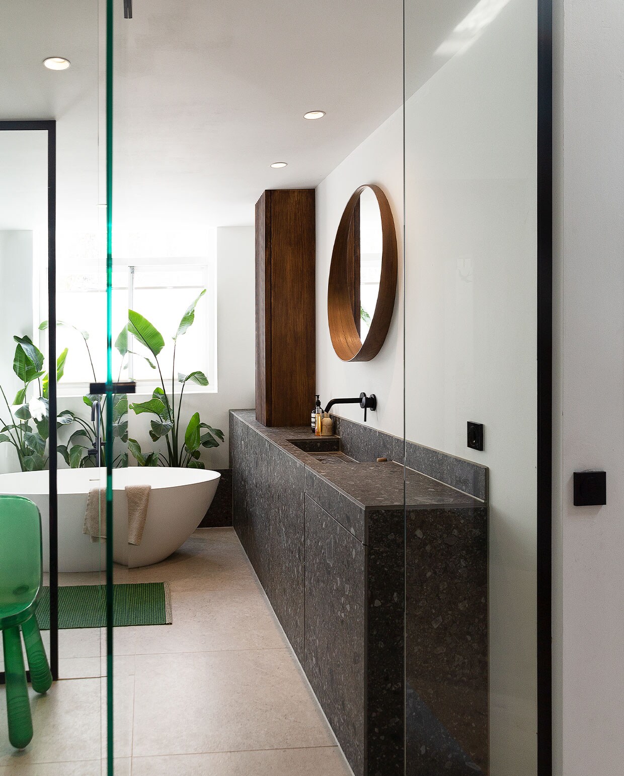 Wastafelmeubel eigen ontwerp, gemaakt van Ceppo di Gré Anthracite van Marazzi. Kraan en badkuip inclusief kraan Best Design. Spiegel Ikea. Hoge smalle walnoot­fineer kast eigen ontwerp. Stoel van Marcel Wanders via Moooi. Badmat Pappelina. Handdoek Zara Home, planten Ikea. Lichtknoppen Berker. Tegelvloer Marazzi, type onbekend. Muurverf Ral 9016 van Fairf. Overig herkomst onbekend.