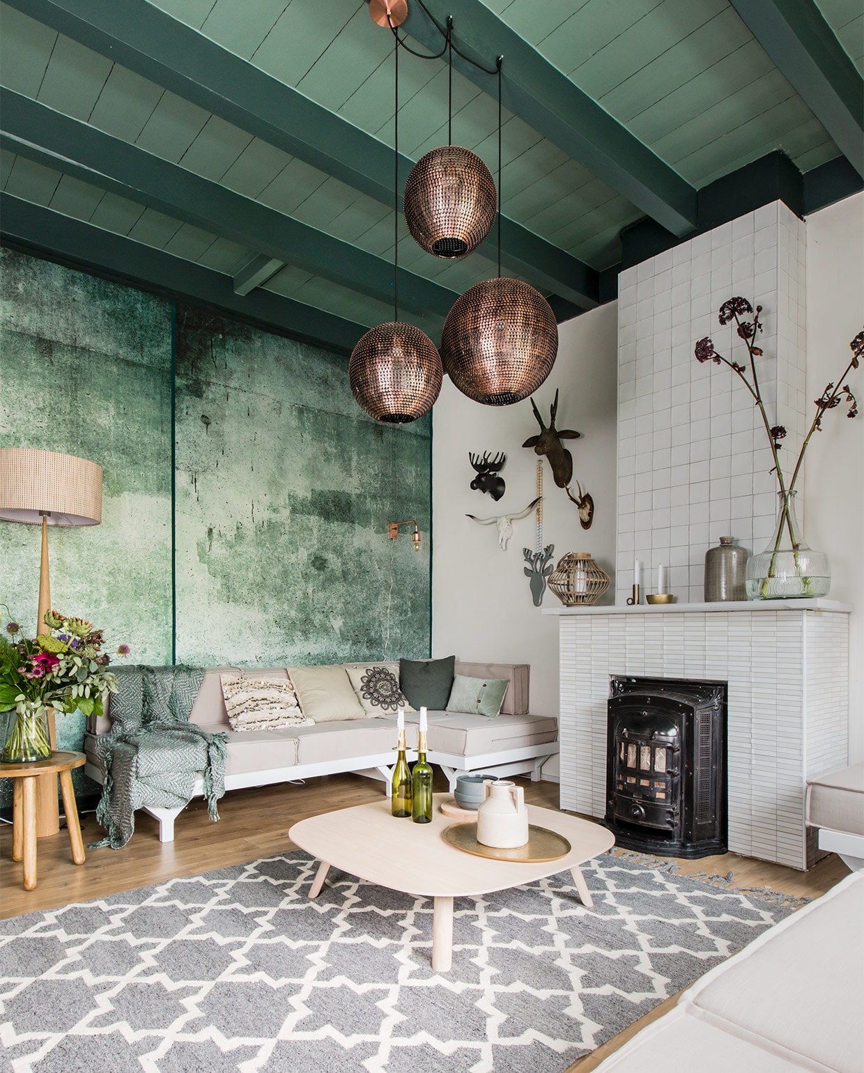 Behang Green Weathered Wall uit de collectie Resource, 4 banen, 212×280 cm (bxh) € 319,- (www.eijffinger.com). Tegels op haard: boven Square Powder wit, 10×10 cm per m² € 64,-, onder Mediterannea Javea Blanco Nieve, 20×20 cm per m² € 51,85 (www.vtwonen.be/vtwonen-collectie). Bank op maat gemaakt, prijs op aanvraag (www.com-anders.nl). Matraskussens soortgelijk, 80×80 cm per stuk vanaf € 115,-, kussen met pailletten € 29,- (www.householdhardware.nl). Vloerkleed 170×240 cm € 299,-, salontafel Tablet, 100x100x25 cm (lxbxh) € 699,-, vloerlamp € 324,95, plaid 180×1340 cm € 64,95, kussen ecru € 29,95, met print € 22,50, groen € 24,95, lichtgroen € 34,95, koperkleurige wandlamp € 119,- (www.eijerkamp.nl). Koperkleurige hanglampen, per stuk vanaf € 119,- (www.dutchbone.com). Bijzettafel Dendon small € 89,- (www.zuiver.com). Hertenkoppen aan wand: wit (linksonder) € 129,-, groot (rechtsboven) € 49,95 (www.vtwonen.nl). Aan ketting € 44,- (www.couleurlocale.eu). Op schouw: windlicht € 29,95, kandelaars € 18,95 en € 24,95, bruine vaas € 39,95 (www.vtwonen.nl). Grote glazen vaas Field Flower € 59,- (www.zuiver.com). Op tafel: grijze pot € 49,- (www.woud.dk). Houten onderbord € 29,95, kruik met oor € 19,95 (www.vtwonen.nl). Goudkleurig dienblad € 79,95 (www.eijerkamp.nl).Overig privébezit.
