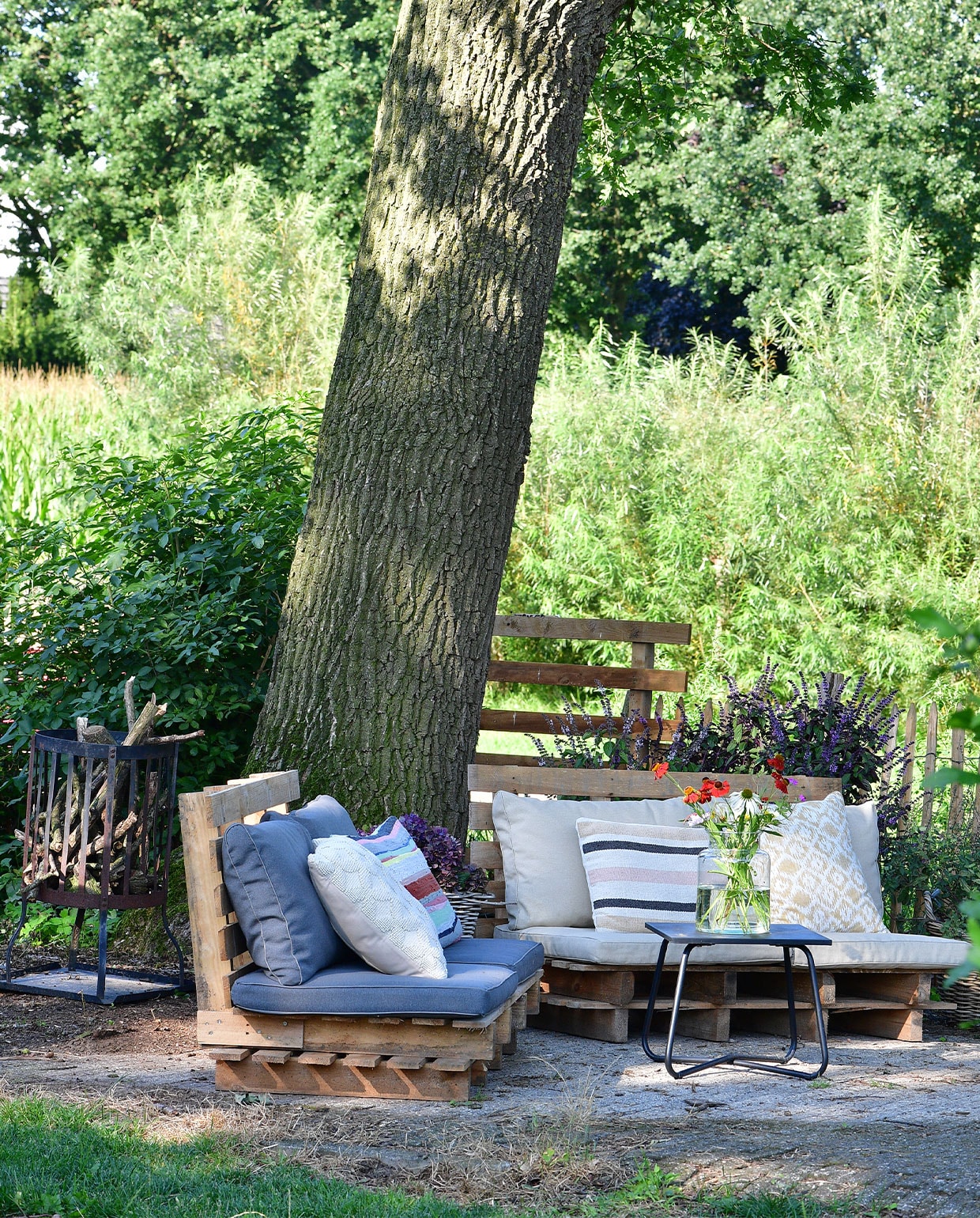 Een fijne zitplek op warme dagen: de zelfgemaakte palletbanken onder de oude eikenboom. Op de achtergrond wilgenbomen. Matraskussens en tafel Kwantum, sierkussens van onder andere fairtrade label Nic &amp; Mic (streep links).
