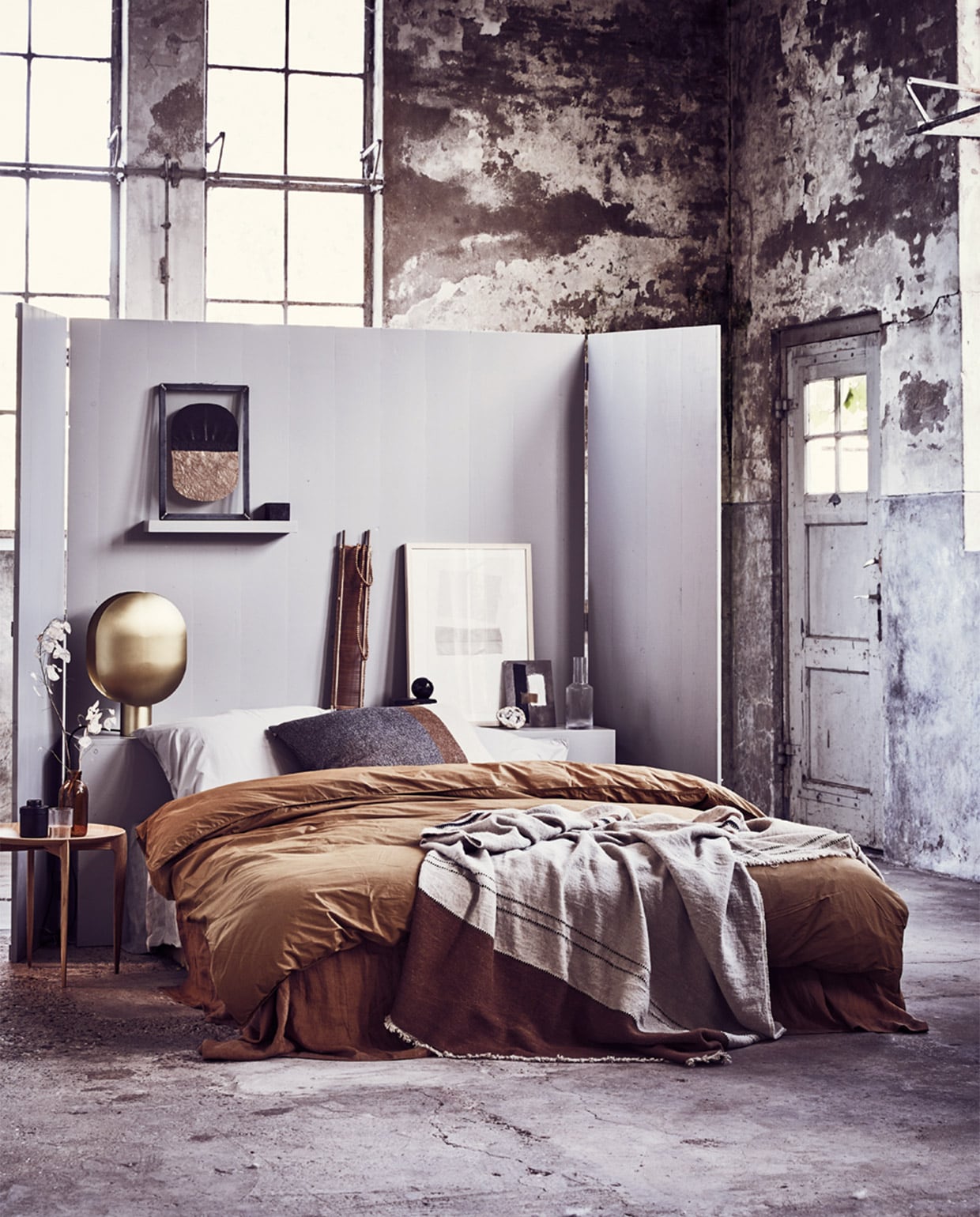 Headboard/kamerscherm (Combitex). Grote lijst met ingelijste textielkunst van Fant, klein kunstwerk op karton van Birgitta de Vos, houten ketting (Sukha Amsterdam). Kunstwerk in stalen frame met papier van Laura de Wilde X Lola Kate 03 (Laura de Wilde). Gestreepte sprei van Libeco van linnen en wol, bruin gestreept kussen 63×63 cm (Couleur Locale). Linnen laken, kaneelkleur, 260×300 cm (By Mölle). Dekbedovertrek perkal katoen, kaki-goud zonder kussenslopen, 220×240 cm, kussenslopen zilvergrijs, 70×60 cm (suite702.com). Glazen karaf, glas van Ferm Living set van 4, zwart potje met deksel (Six and Sons). Salontafel Tray Table van Fritz Hansen (Mobilia). Messing lamp Clamp van Norr11 (Melchior Interieur). Smalle houten zeef (Aura Peeperkorn). Overig privébezit.