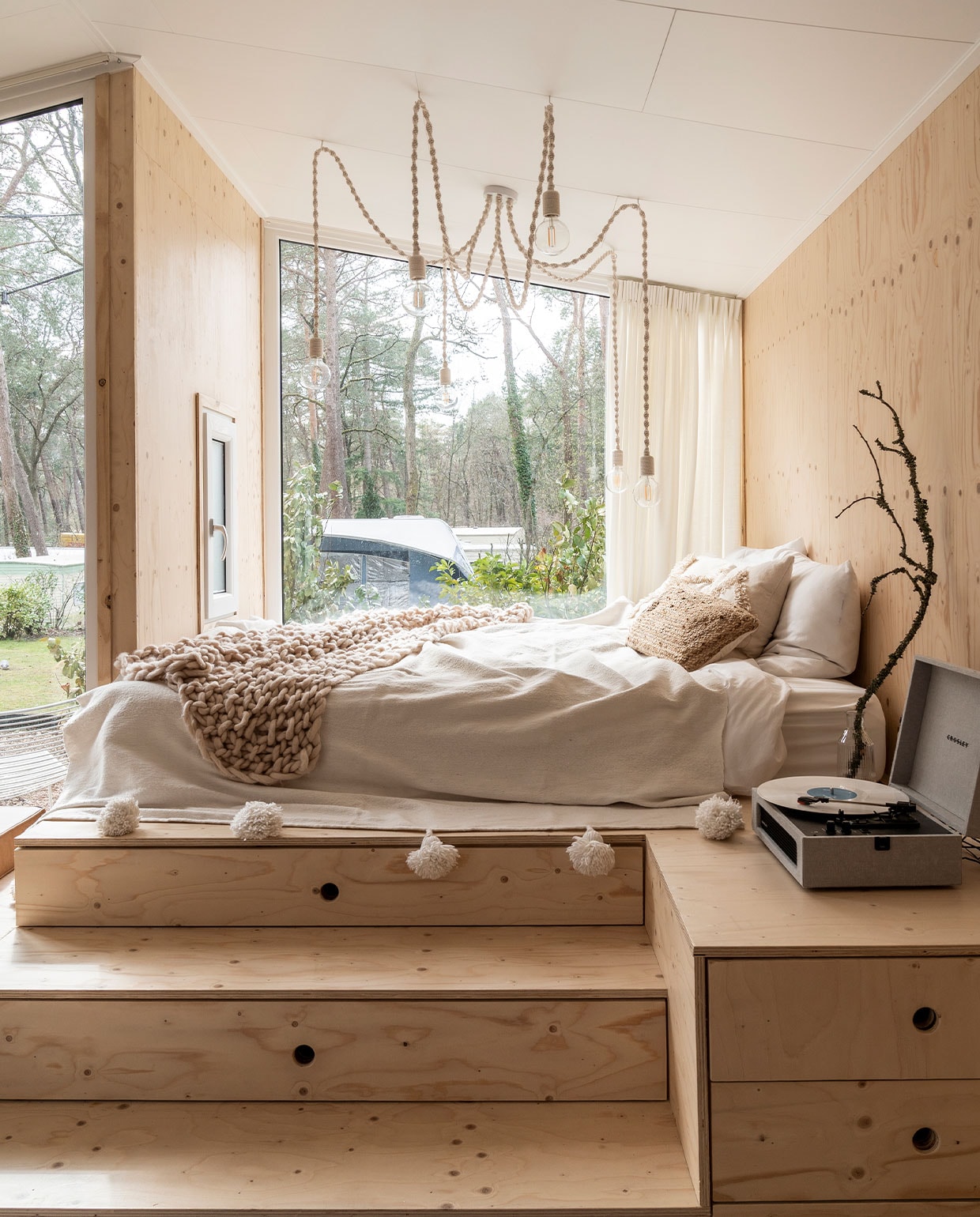 Bedombouw Hertog Recreatiebouw.Kandelaarlamp zelf­gemaakt. Matras Emma, beddengoed herkomst onbekend, sprei met pompons gekocht in Marrakech, gehaakte sprei Lidl. Waterkaraf Ikea, tak uit het bos, platenspeler Crosley, beige kussen H&amp;M Home, bruin kussen Calma House.