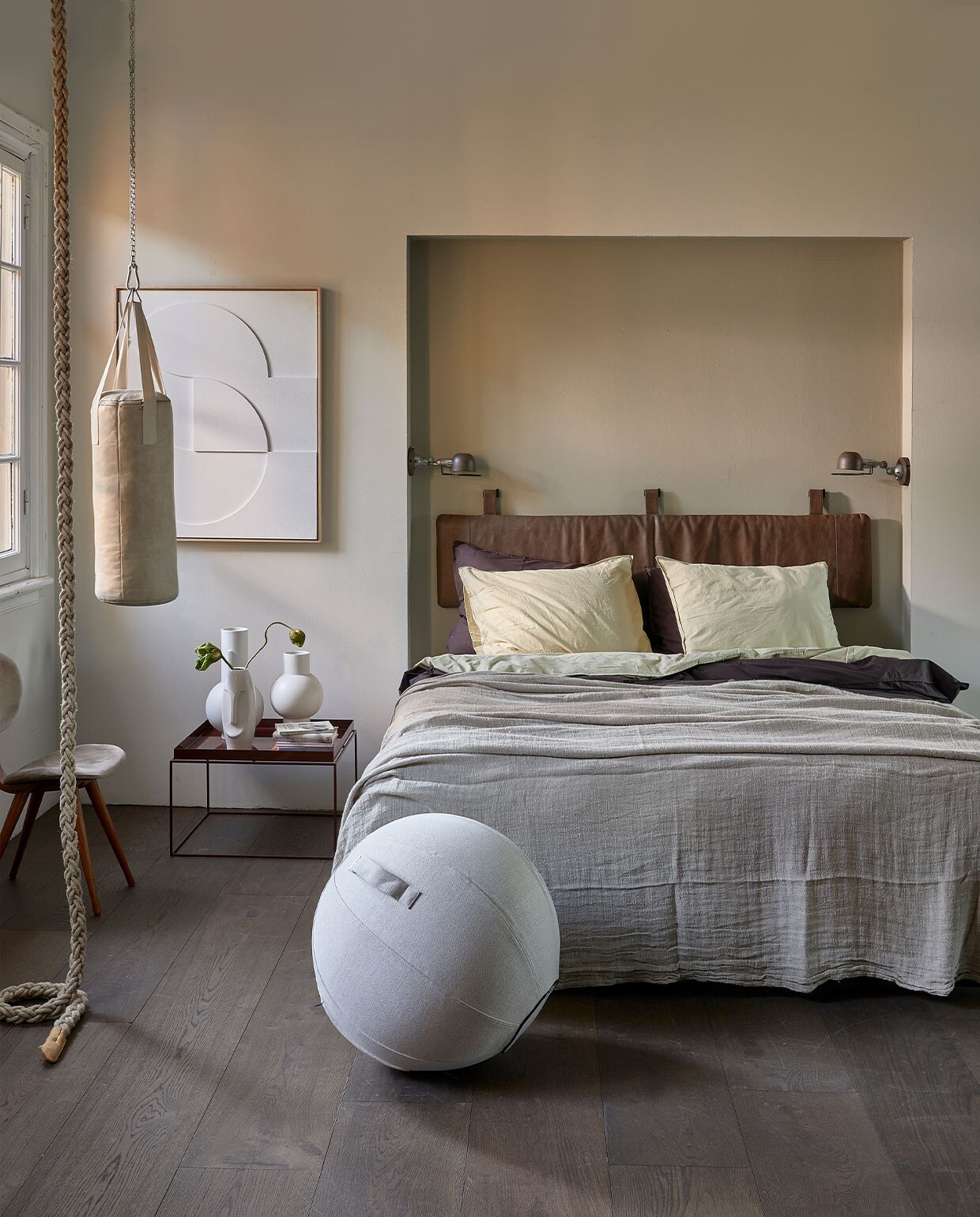 Muurverf krijtlook mat van vtwonen, kleur achter bed Soap, eromheen Linen, per liter € 14,20, vloer laminaat XL, kleur board per m2 € 25,99 (karwei.nl). Hoofdbordkussen met lus en beugel, 40×180 cm € 339,- (nahka.nl). Boxspring Espevar, 180×200 cm € 858,-, dekbed Smasporre, 240×220 cm € 29,99, hoofdkussens Gronamarant, 50×60 cm, per stuk € 19,99, hoeslaken Dvala, 180×200 cm € 29,99, beige/groen katoenen dekbedovertrek en slopen Angslilja, 240×220 cm € 34,99 (ikea.nl). Linnen beddensprei naturel, 270×270 cm € 229,-, matwitte vazen achterste op tafel, 21×32 cm (Øxh) € 42,95, vaas daarnaast 21×42,5 cm (Øxh) € 49,99, relief art paneel € 595,- (hkliving.nl). Veganleren boksbal € 549,- (sukha.nl). Klimtouw € 75,-, stoel Pastoe met fluwelen bekleding € 295,- (neeflouis.nl). Donkerbruin katoenen dekbedovertrek Ambiante, 200×220 cm € 46,25, wandlampen Lucide Honore, per stuk € 55,99, zitbal Vluv Sova, 65 cm (Ø) € 105,-, vaas Erla met gezicht, 31 cm (h) € 24,95, bijzettafel Tray van Hay, kleur chocolate € 228,- (vtwonen.be). Reliëfkunstpaneel € 625,- (hkliving.nl).