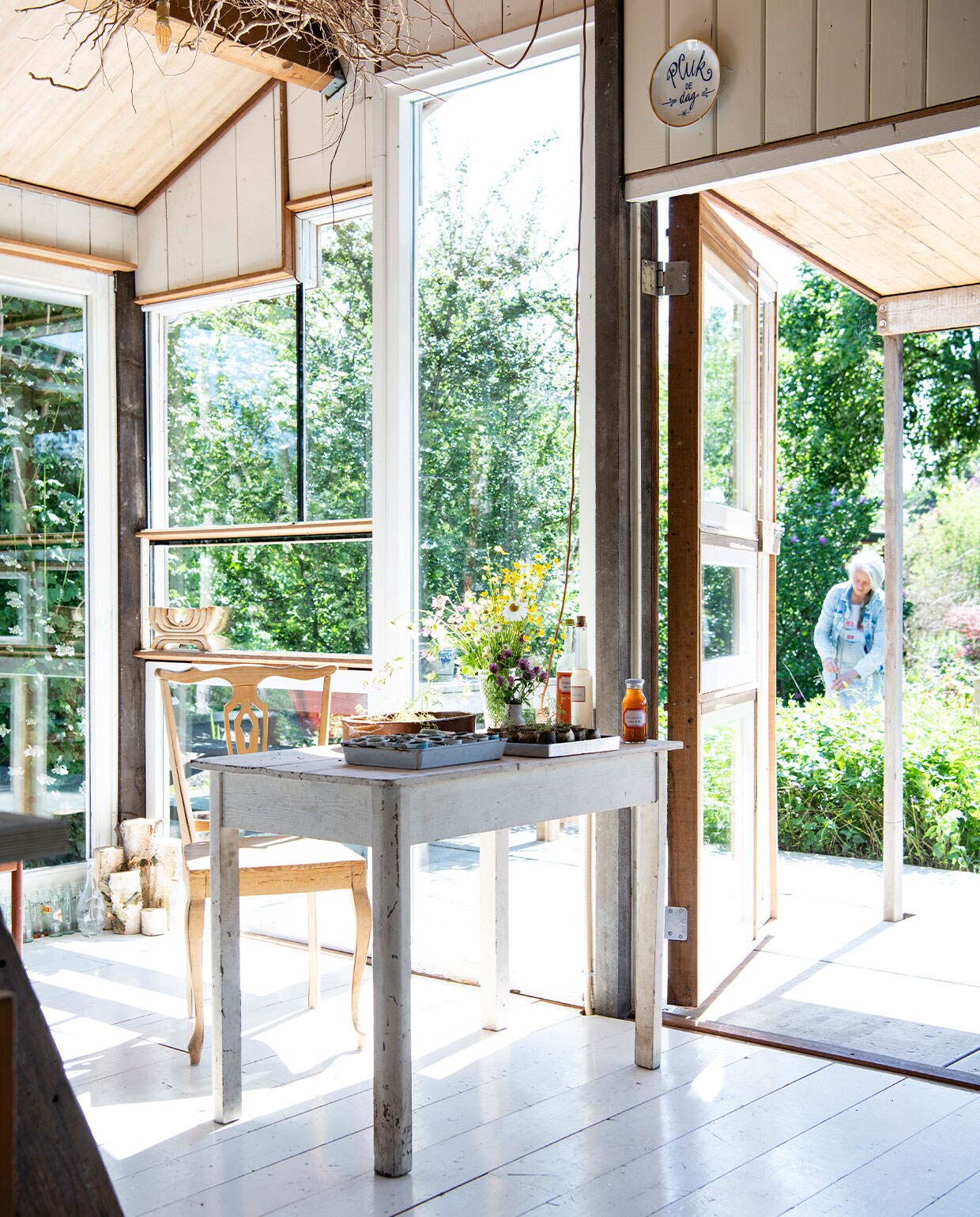 Witte vloerplanken gekregen van een vriendin, deuren en ramen gemaakt van kozijnen gevonden in containers, bord boven deur gemaakt door Simone te koop in haar webwinkel winkelgloria.nl. Tafel kofferbakverkoop Buitenhuis, stoel kringloop, bakje op tafel Old Harry’s, houten kandelaar in raam kringloop Vintage &amp; More. Flessen met zelfgemaakte vlierbloesem- en seringensiroop. Veldboeket met bloemen uit eigen tuin.