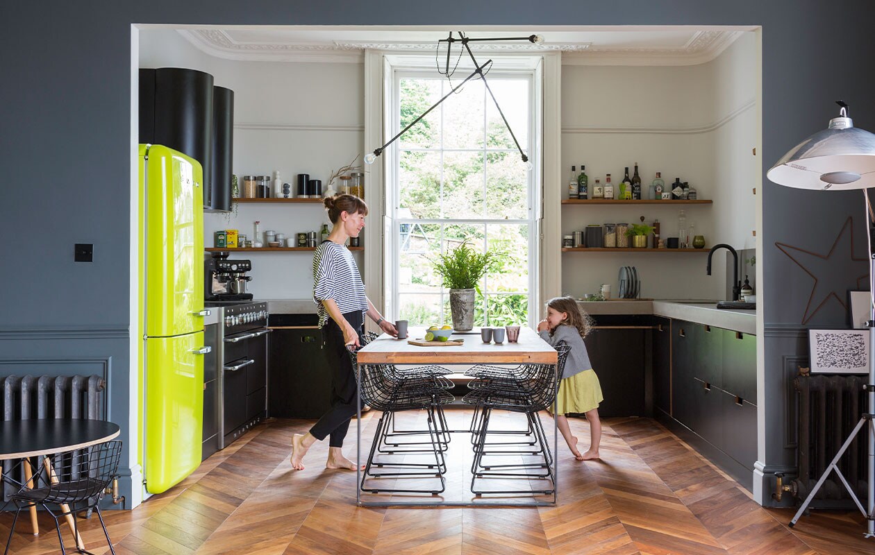 Muurverf Matt Emulsion in kleur French Grey Pale van Little Greene. Koelkast Smeg. Eettafel gemaakt van een oude kerkdeur, vintage stoelen Bertoia, kroonluchter zelfgemaakt. Keukenkasten Ikea met op maat gemaakte frontjes en deuren van Matt Antrobus, werkblad Mass Concrete, afzuigkap CDA, planken gemaakt van gebruikt hout, kraan Franke. Espressomachine Sage. Voorraadpotten Orla Kiely. Vloer Hicraft. Onderstel tafel Stone Butterfly met zelfgemaakt blad van multiplex. Overig onbekend.