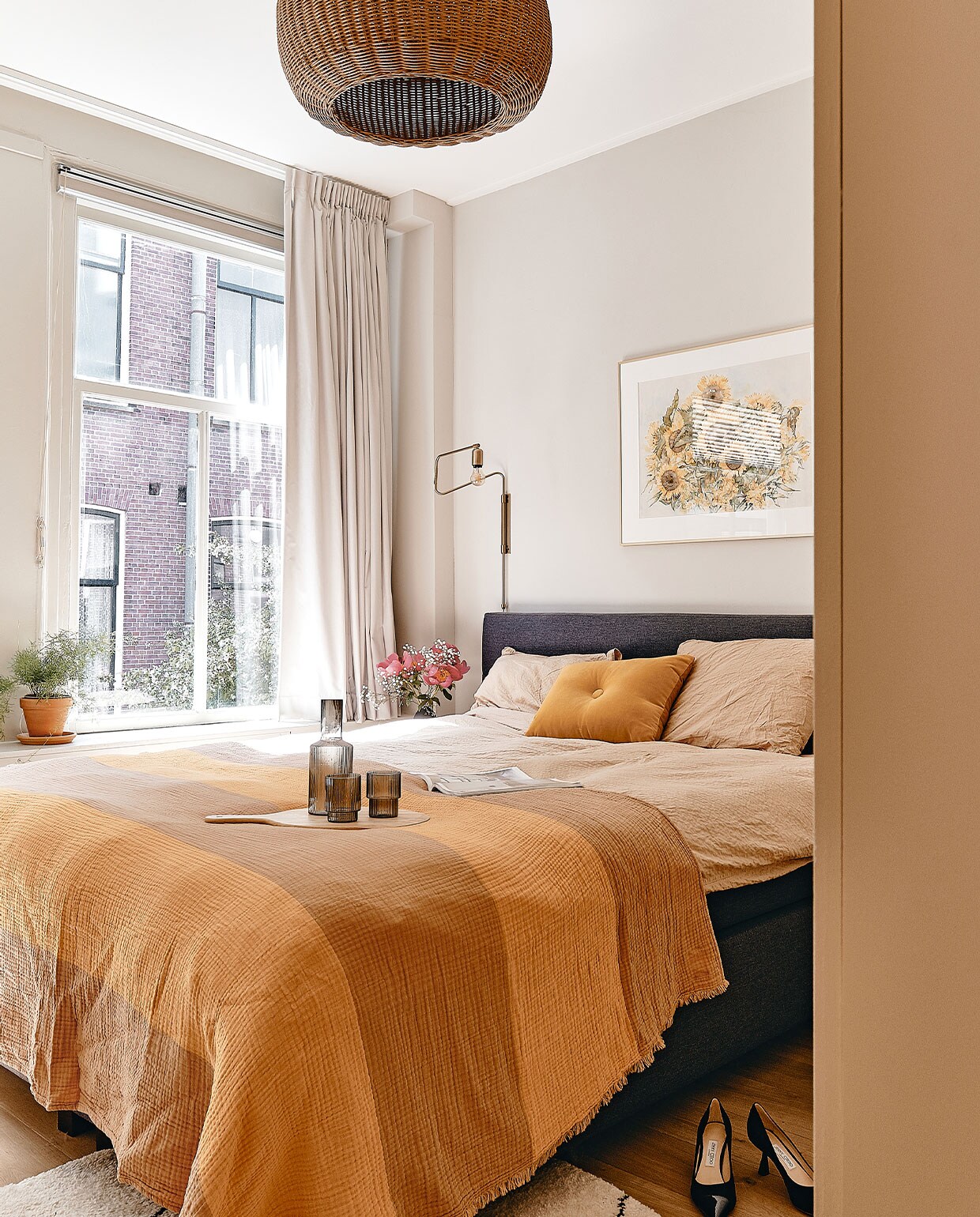 Muurverf Ammonite van Farrow &amp; Ball. Wandlamp Swing van House Doctor. Hanglamp Braided Belly van Ferm Living. Ingelijste zonnebloemen van Maartens ouders. Beddengoed Crisp Sheets, oranje kussen en sprei Hay. Dienblad Ikea, karaf en glazen Ferm Living. Vloerkleed Benuta, pumps Jimmy Choo. Kast Pax van Ikea, geverfd in Hay van Farrow &amp; Ball.