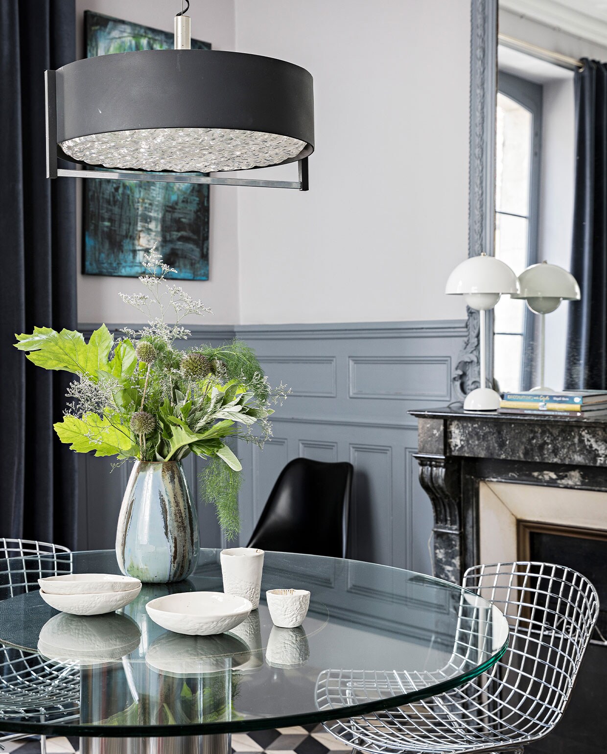 Vintage eettafel van glas en metaal uit 1970, vintage Wire Chairs van Harry Bertoia, jaren 70 hanglamp van onbekende makelij, serviesgoed van Myriam Aït Amar Ceramics, kunstwerk acryl op canvas van Clarisse Friedmann, op de schoorsteen Flowerpot lamp van Verner Panton. De cement- tegels zijn origineel, muurverf van Farrow &amp; Ball.