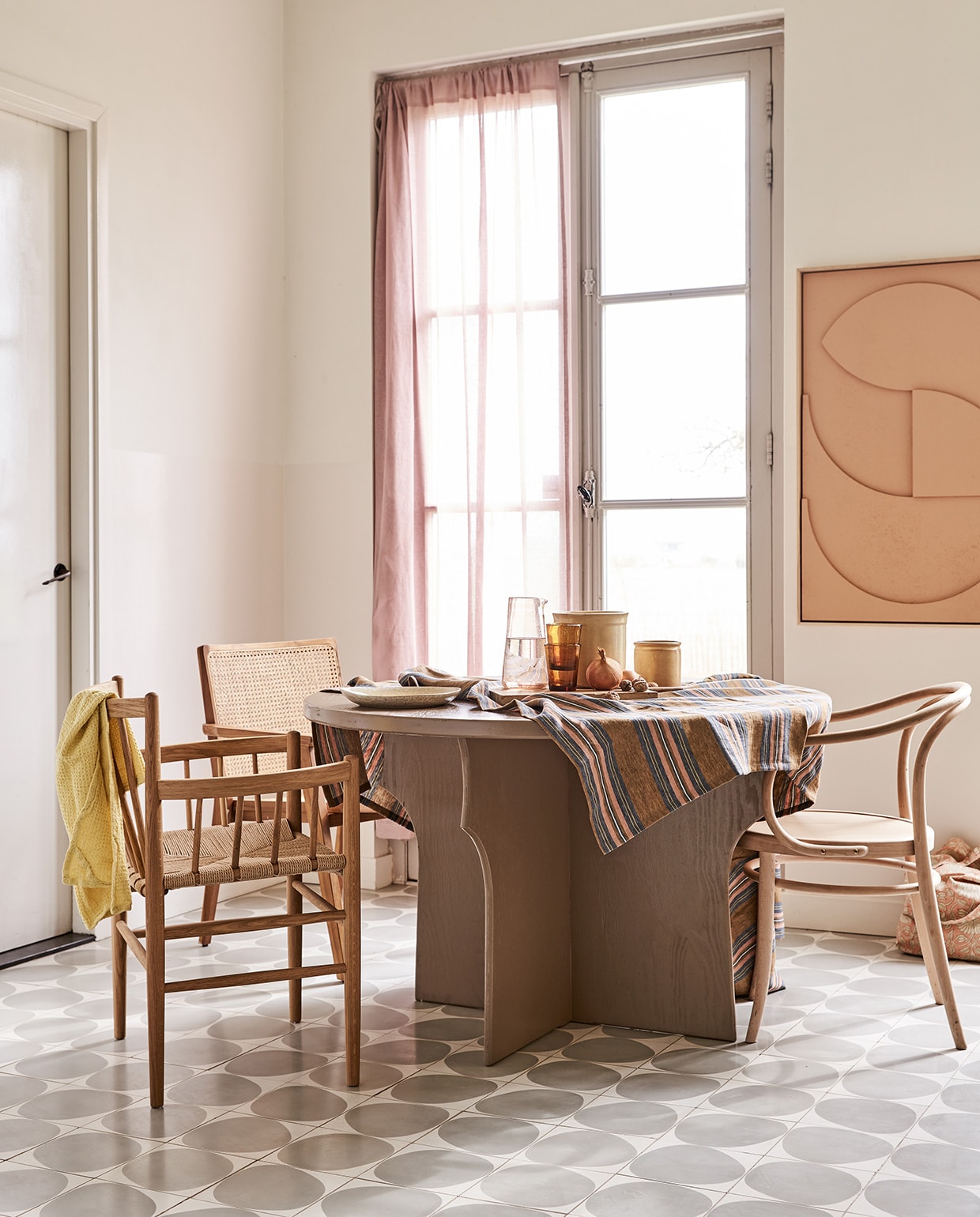 Tafel gemaakt naar eigen ontwerp en geschilderd in kleur Mexican Sand, prijs op aanvraag (com-anders.nl). Stoel met spijlen FD Møbler J81 Jørgen Baekmark (letoile.store). Stoel met webbing zitting Nova (vtwonen.nl). Stoel Thonet-model (dille-kamille.nl). Tafelkleed (Libeco). Karaf, water- glazen Salon Amber, bord, theedoek wafel in kleur Lemon, tas met roze bloemen (scandicliving.nl). Artwork aan de wand in kleur perzik, 60×80 cm (hkliving.nl). Gordijn van voile stof Cosia in de kleur Orchid van Designers Guild (wva.nl).