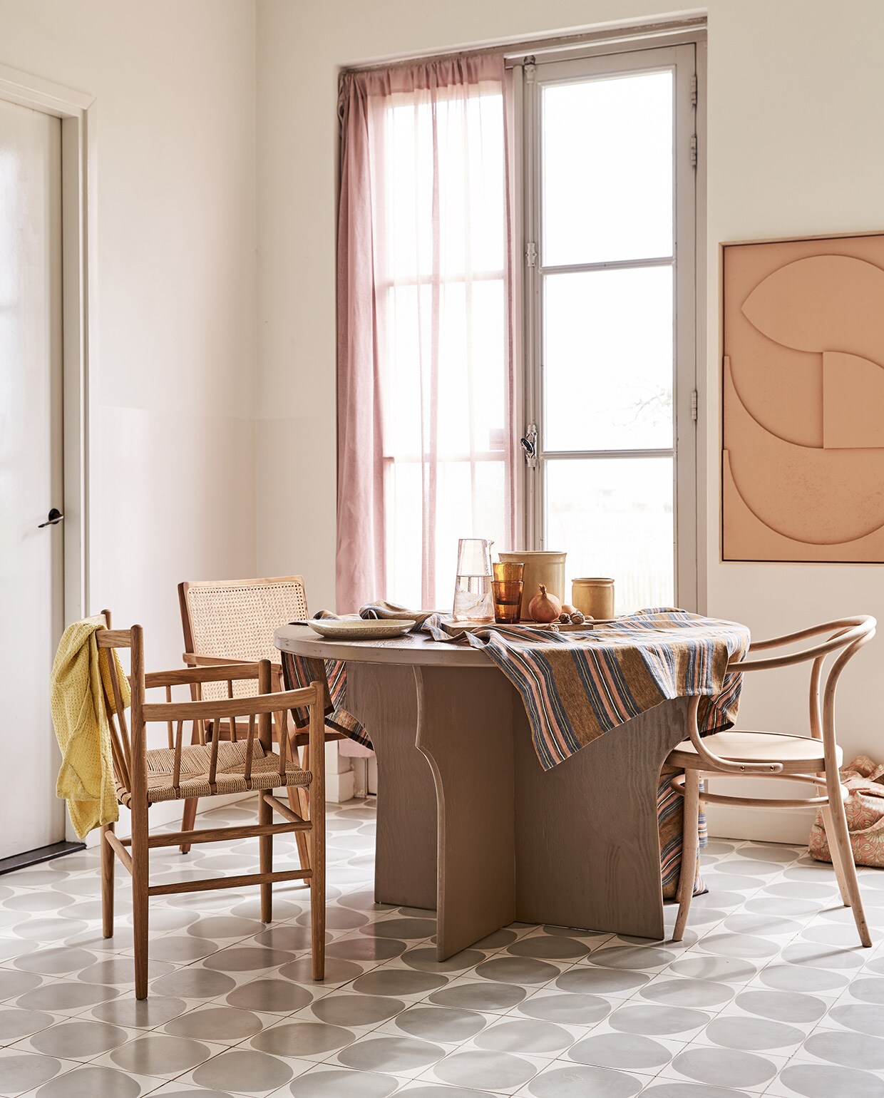 Tafel gemaakt naar eigen ontwerp en geschilderd in kleur Mexican Sand, prijs op aanvraag (com-anders.nl). Stoel met spijlen FD Møbler J81 Jørgen Baekmark (letoile.store). Stoel met webbing zitting Nova (vtwonen.nl). Stoel Thonet-model (dille-kamille.nl). Tafelkleed (Libeco). Karaf, water- glazen Salon Amber, bord, theedoek wafel in kleur Lemon, tas met roze bloemen (scandicliving.nl). Artwork aan de wand in kleur perzik, 60×80 cm (hkliving.nl). Gordijn van voile stof Cosia in de kleur Orchid van Designers Guild (wva.nl).