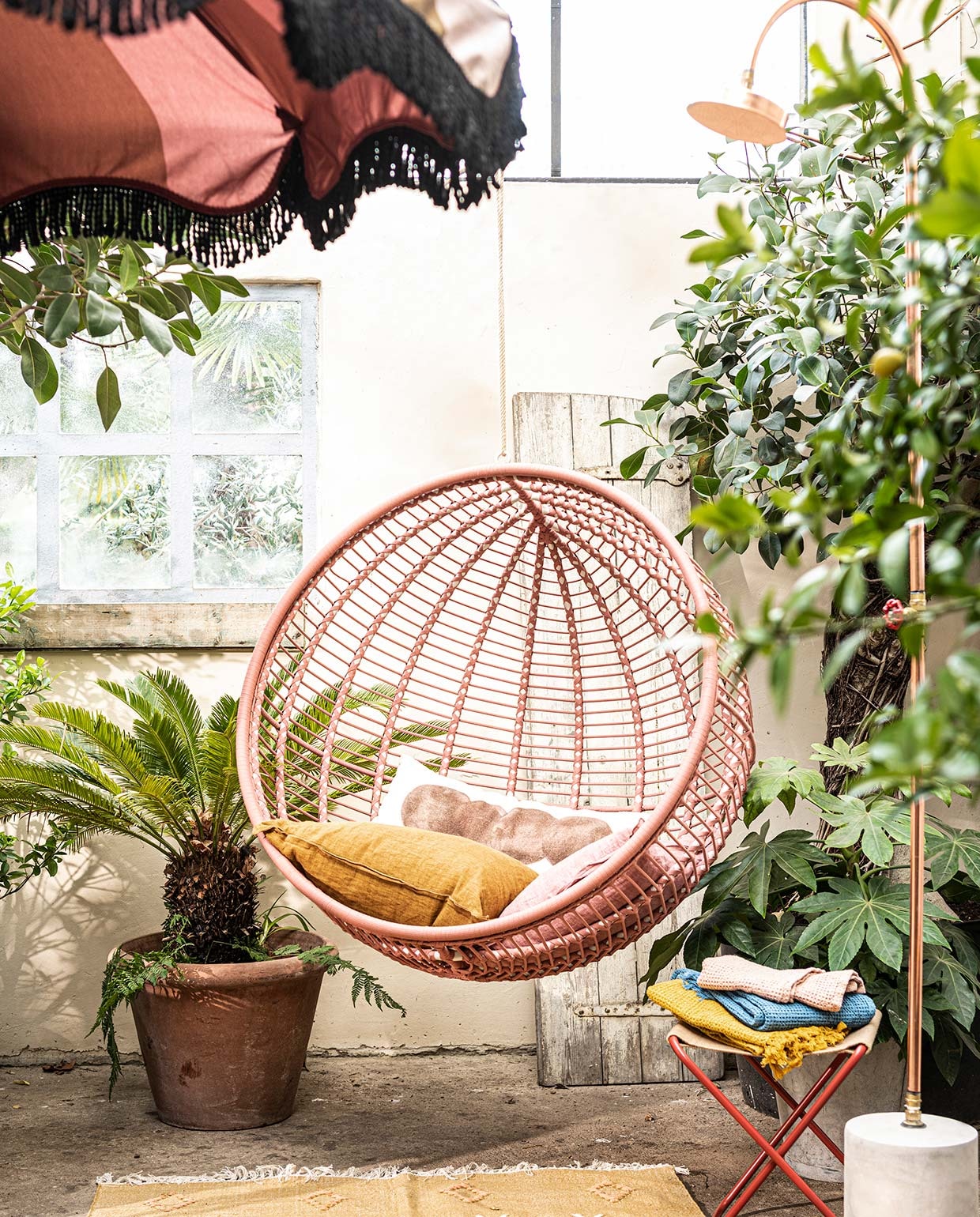 Stalen hangstoel voor buiten in zacht terra, 107×73 cm (Øxd)(kavehome). Krukje Desert van Ferm Living (vtwonen.be). Koperen buitendouche van Seletti (vtwonen.be). Parasol van HKliving (vtwonen.be). Langwerpig gestipt kussen (urbannatureculture.com ). Linnen kussens, wafeldoeken (bungalow.dk). Vloerkleed van Madam Stoltz (deens). Plant en pot privébezit.