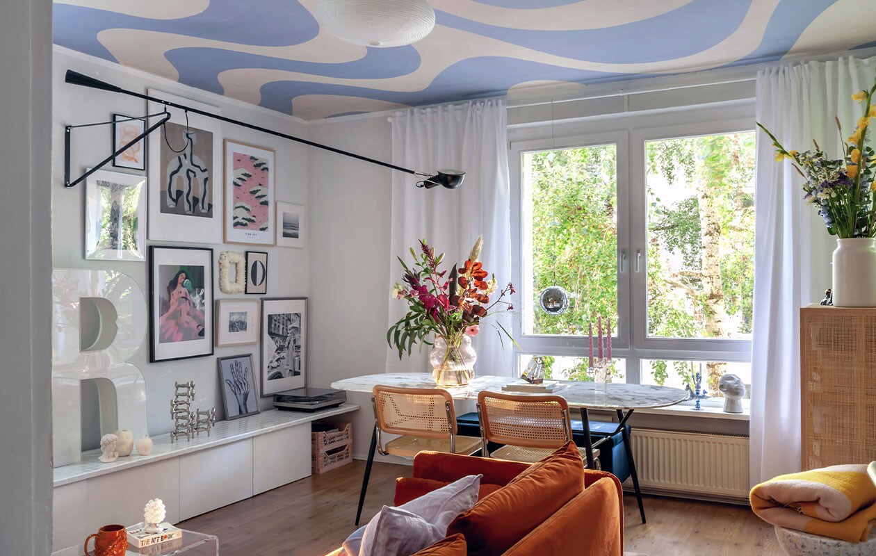 Witte lage kast (met zelf betegeld blad) en houten kast Ikea, R lichtletter via Ebay Kleinanzeigen, tafel Depot, koffietafel via Ebay, stoelen Home24, lamp Flos, beker zelfgemaakt, kruk Wild Studio, plaid Arket, glazen vaas Pols Potten, kaars Sentimental Candles. Behang op plafond Curvy van Belarte Studio.