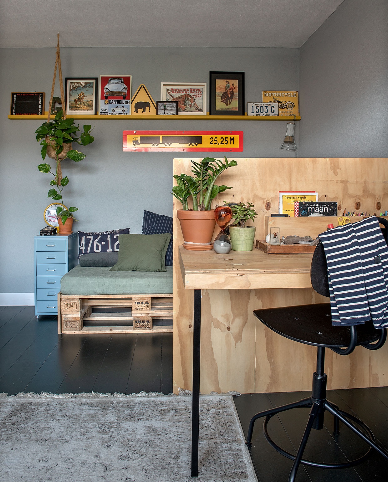 Verf vloer Kaviaar van Histor, wand Pastelblauw van Karwei. Chillbank gemaakt van pallets en een matras bekleed met een deken van Kwantum. Kleed Karwei, bureaustoel, ladekast, klemlamp Ikea. Nummerbord USA, klok Loods 5. Zandloper Leen Bakker. Plantenpotten Intratuin. Op de wandplank pagina’s gescheurd uit een oud circusboek. Langwerpige vrachtwagen via Bas’ werk. Overig onbekend.