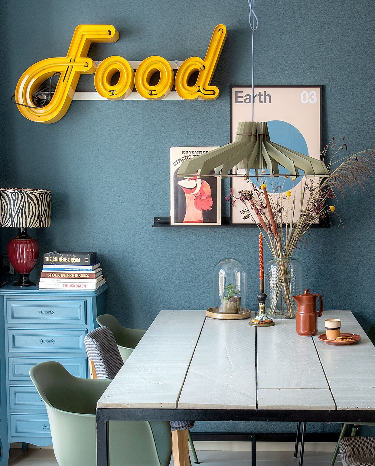 Wandkleur Vintage blauw van Karwei. Vintage neon gekocht van een bevriende leverancier. Tafel zelf gemaakt, kuipstoelen Karwei, grijze stoel Joe van Midj. Hanglamp Van Tjalle &amp; Jasper. Lampenkap Kwantum, vaas Leen Bakker. Poster Earth van Desenio, ernaast een oud circusboek. Stolp, bord Mica Decorations. Beker HKliving. Kast, lampvoet, koffiekan, kandelaar van de kringloop. Stalen schilderijrichel gemaakt door Bas en collega.
