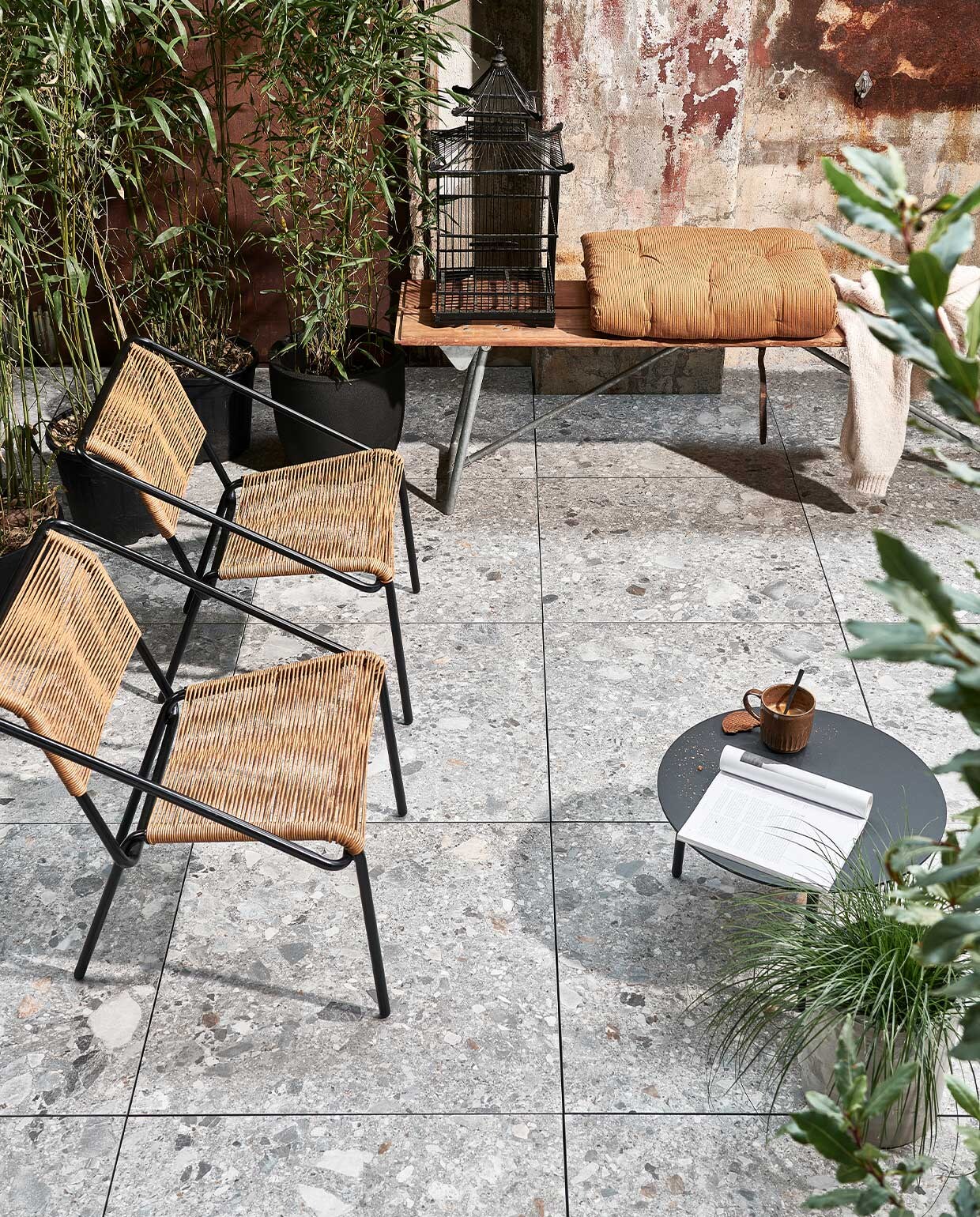 Buitentegels Solostone Uni Composite mix, 70x70x3,2 cm per m² € 69,95. M.m.v. basiclabel.nl (fauteuils Wisp van BePureHome), com-anders.nl (bank, vogelkooi), vtwonen.nl (bijzettafel van Serax, beker, grijze betonlook bloempot), deens.nl (terrakleurige matras van Madam Stoltz).