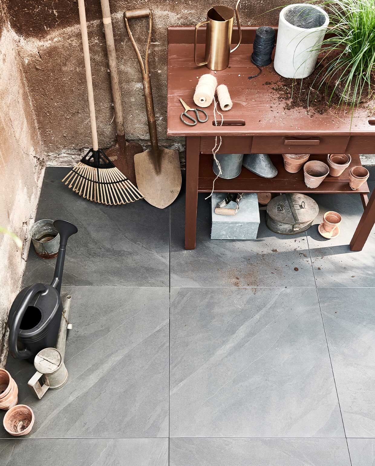 Buitentegels Solostone Uni Waves Antracite, 70x70x3,2 cm per m² € 69,95. M.m.v. vtwonen.nl (klapkruk met stoffen zitting van Ferm Living, zwarte gieter van Elho), com-anders.nl (scheppen, houten tafel, oud zink, mand met strepen), intratuin.nl (terracotta potten, hark van hout en bamboe, snoeischaar).