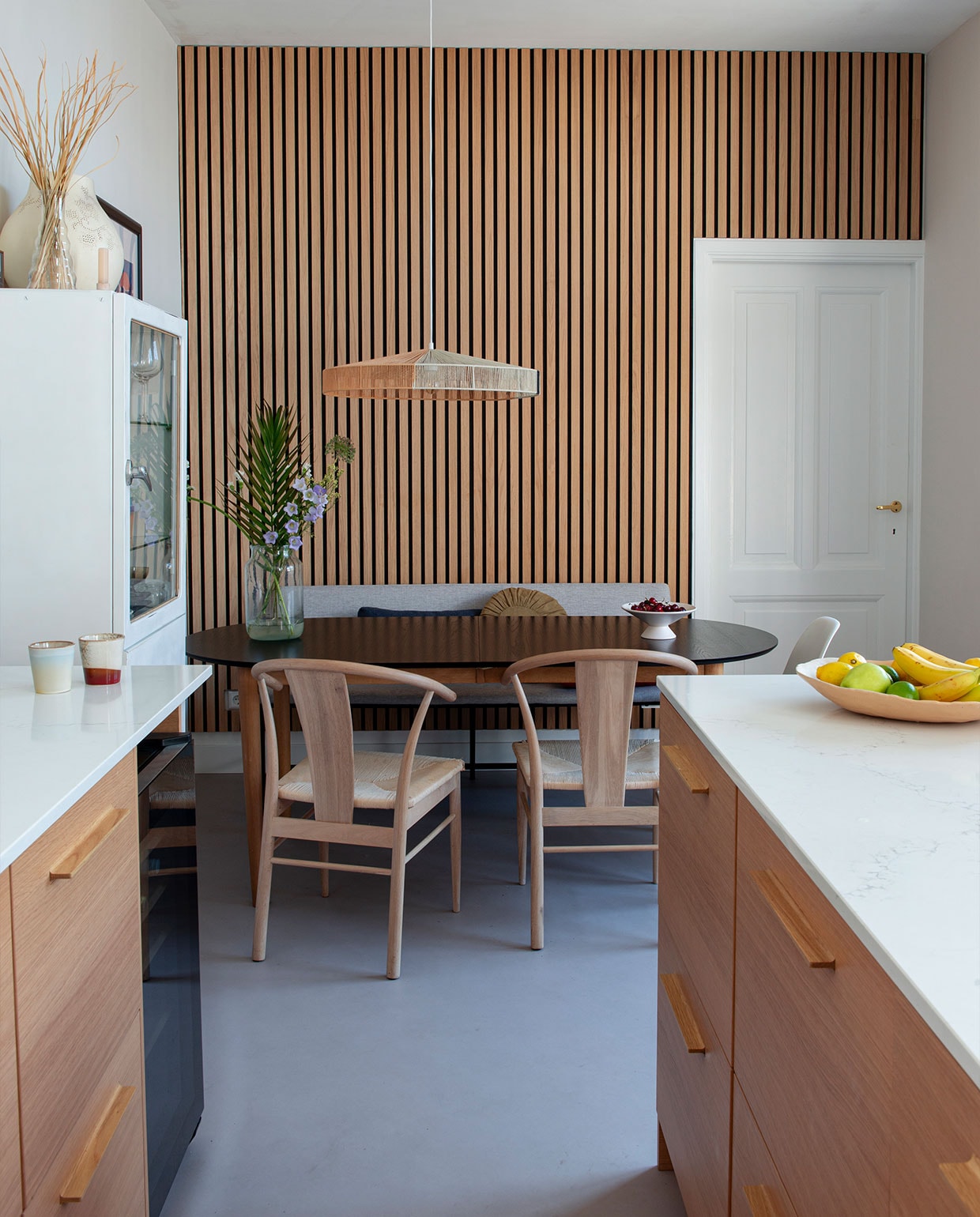 Keuken Kvik met composietblad in marmerlook, akoestische eikenhouten wandpanelen en houten stoelen Lijn M, eettafel Jysk, hanglamp HKliving, vintage apothekerskast uit Polen, bankje Wehkamp.