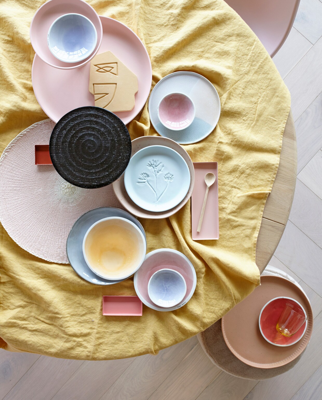 Parket visgraat eiken wit geolied van vtwonen (karwei). Ronde eikenhouten eettafel, roze eetkamerstoel (coosdewitwonen). Geel linnen tafelkleed (letoile.store). Roze etagère (loods5.nl/design). Schaaltjes XS in blauw en roze, S in blauw, M in geel, roze en rood (studioharmenelke). Ornament gezicht, rond bord met opstaande rand, rond terra dienblad (hkliving). Zwarte houten etagère (paviljoennoord.com). Roze placemat (klevering). Niet perfect rond bord met spikkeltjes, vergrijsd blauw bord (loods5). Lichtblauw bord van keramiek met bloemprint (letoile.store). Rechthoekige schaaltjes in verschillende tinten roze, geel glas (hm.com). Houten lepel (restored). Roze fluwelen poef (covanderhorst).