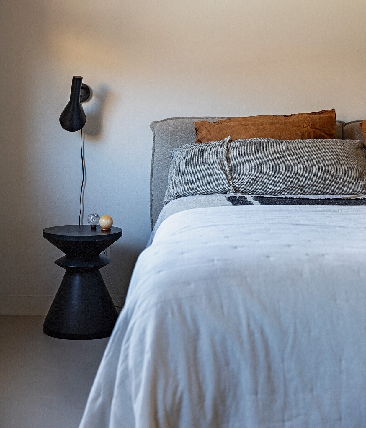 Muurverf Papyrus van Gamma, bed en bijzettafel La Redoute. Wandlamp AJ vanLouis Poulsen, item op bijzettafel erfstuk van Ediths vader. Wit, lichtgrijs enkaneelkleurig beddengoed H&amp;M Home, grijs beddengoed Zara Home, grijsblauwbeddengoed met streep The Recollection.