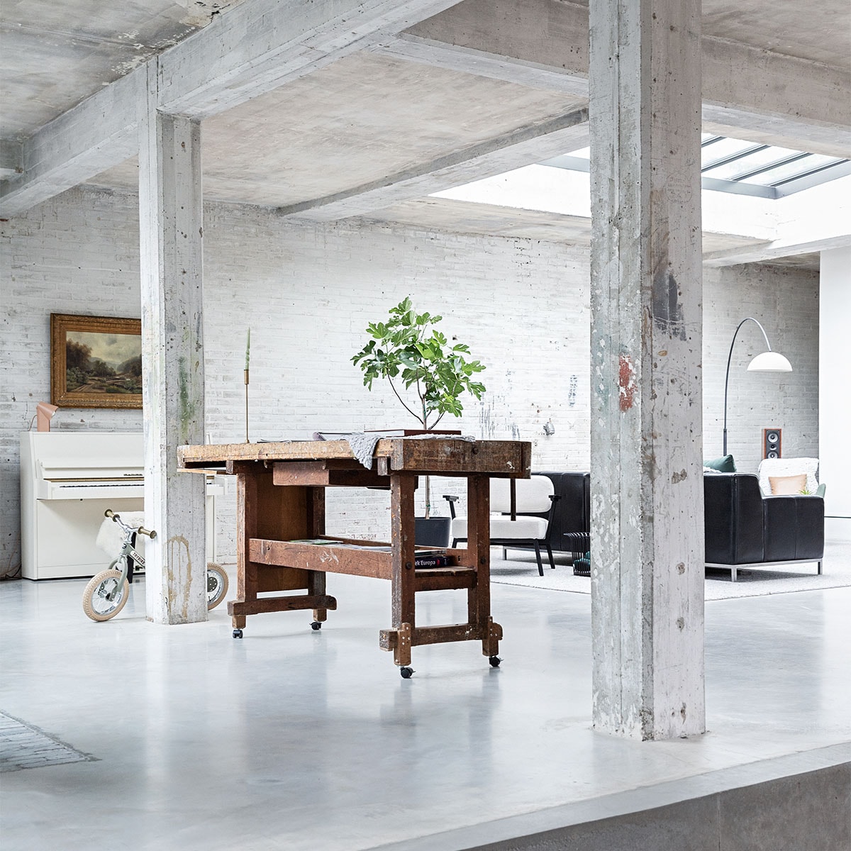 Vloer gevlinderd beton. Hardstenen waaltjes Spotted Bluestone. Werkbank erfstuk meubelfabriek. Kinderfiets Trybike. Witte fauteuil Ode Lounge Chair van Studio Henk.