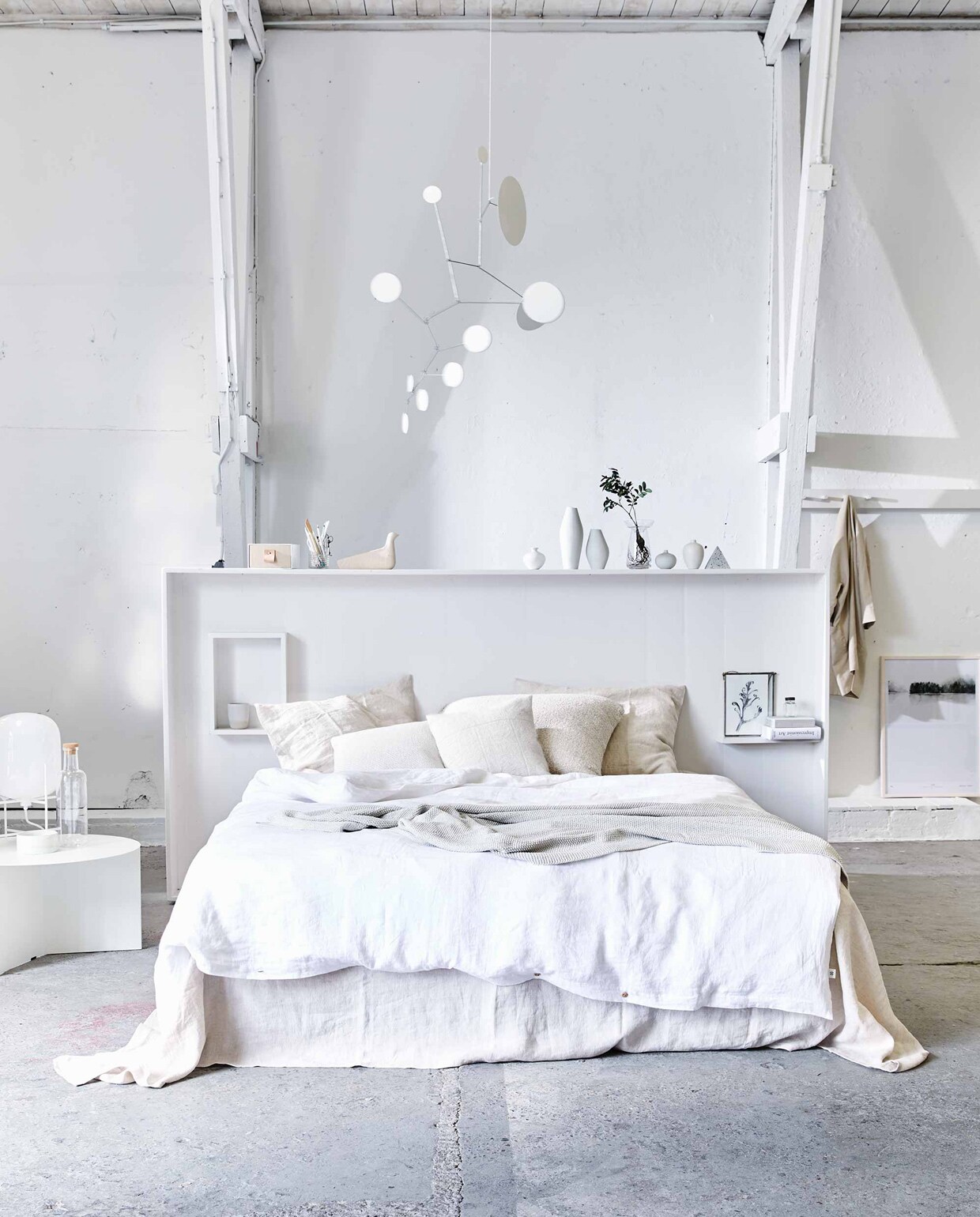 Hoofdbord (Combitex). Linnen laken Almond, linnen dekbedovertrek Pure white, kussenslopen Seashell, gerecyclede denim plaid in off white (By Mölle). Bijzettafel Slit Table XL White, fles met kurken dop (Hay Amsterdam). Lamp Pulpo small white (Mobilia). Kleine vaasjes (Maison NL). Hoge vazen van porselein (CK Ceramics). Piramide van steen (Restored). Glazen bollenvaas, linnen jas (Six and Sons). Afbeelding Calm Botanics in lijst (My Deer’s). Witte dot mobile (Lappalainen via Etsy). Lage schaal op tafel, beker in kast (Annemieke Boots Ceramics). Opbergdoos met nude laatje (&amp; Other Stories). Kussen van witte alpaca wol, groot kussen Colombia (Couleur Locale). Witte linnen kussenhoes (H&amp;M Home). Houten Eames vogel (Homestede). Tekening in lijst van Anke Loijens en overig privébezit.