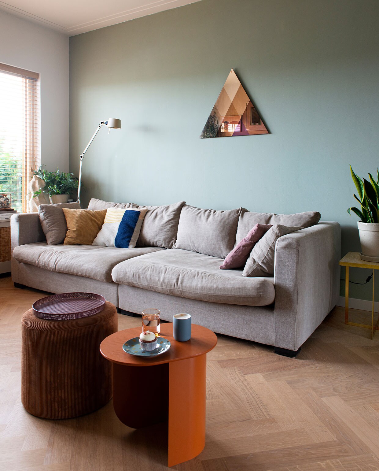 Linnen bank Chuck van Homestock, kussens op bank Ferm Living. Kleur achter bank Smoking Grass 23 van Vestingh Paint. Driehoekige spiegel Transcience van Lex Pott. Terrazzo bijzettafel met goudkleurig onderstel Luigi van Zuiver. Bijzettafel Plateau van Fest. Poef en dienblad Karwei. Bord Hema, mok Archive Studio, glas &amp;klevering. Vloerlamp Bolt van Tonone. Overig onbekend.