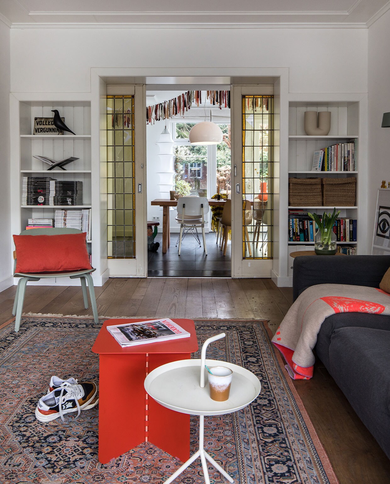 Vloer van Amerikaans eiken lag er al. Muurverf wit RAL 9016 Sikkens. Grijze bank gekocht tijdens een garagesale. Plaid op bank Home Sweet Home Adelheid Laden.Vloerkleed kringloop. Sneakers Atlantic Stars. Rode bijzettafel Puik Hinge. Witte bijzettafel Hay. Mok HKliving. Links in kast: drankvergunning uit café van vader, houten kraai Eames House Bird Vitra, boeken­standaard Mendo. Groene Lounge Chair Muuto. Rood kussen Kus-sen. Rechts in kast: vaas Kink Muuto, manden Ikea. Bruine bijzettafel Kwantum. Erop vaas Muuto. Schuifdeuren Marktplaats.