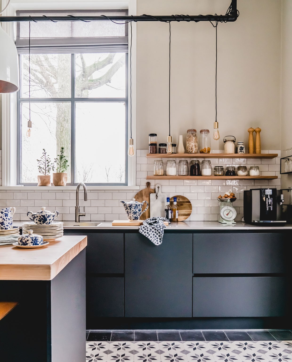 Ook de keuken kreeg nieuwe Portugese cementtegels van Floorz. De keuken is van Tulp. De reling met lampen maakten ze zelf van steigerpijp (steigerpijp.nl) en strijkijzersnoer (snoerboer.nl). De lamp boven de bar, waar ze graag aan ontbijten, is van Pols Potten. Het grijze vouwgordijn is van maatstudio.nl. Het blauwe servies komt uit de familie van Jankees, de voorraadpotten van Marjoleins opa. Plantenzakjes van Uashmama.