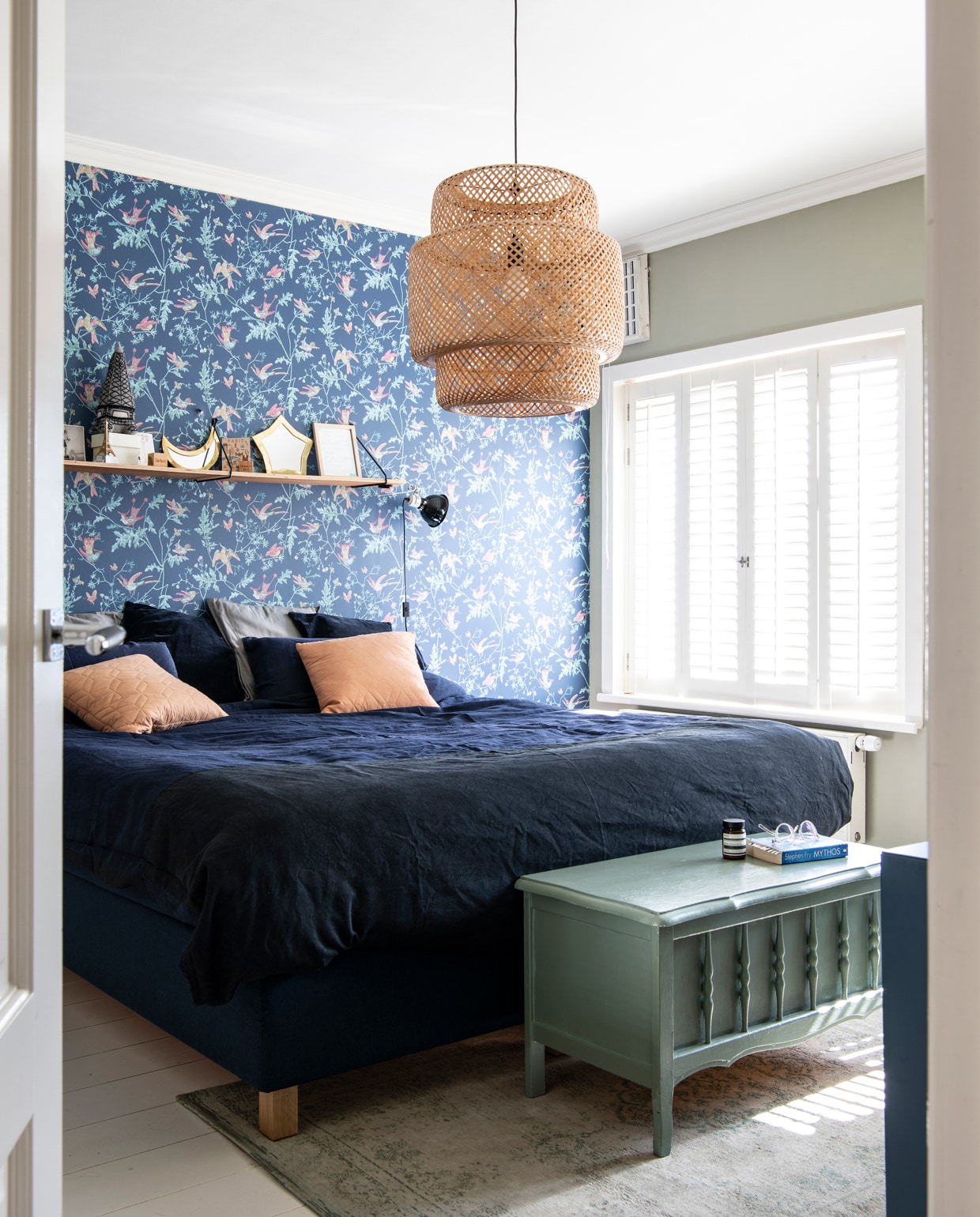 Behang Hummingbirds van Cole &amp; Son via Co van der Horst. Wandplank met houders House Doctor. Op de plank: maan en ster spiegels The Souks, Eiffeltoren-knuffel Oeuf NYC, doos Hay, houten blokken met gelaserde afbeelding geboortekaartjes kinderen Uncover Lab. Bed Eastborn van Bedvisie, blauw beddengoed Crisp Sheets. Terra kussen Fest Amsterdam, blauw velours kussen en rotan lamp Ikea. Vintage dekenkist familiebezit. Vloerkleed van Louis de Poortere via Felice. Muurverf French Gray van Farrow &amp; Ball. Vloerverf Soft Pearl van Flexa. De raambekleding zat al in het huis.