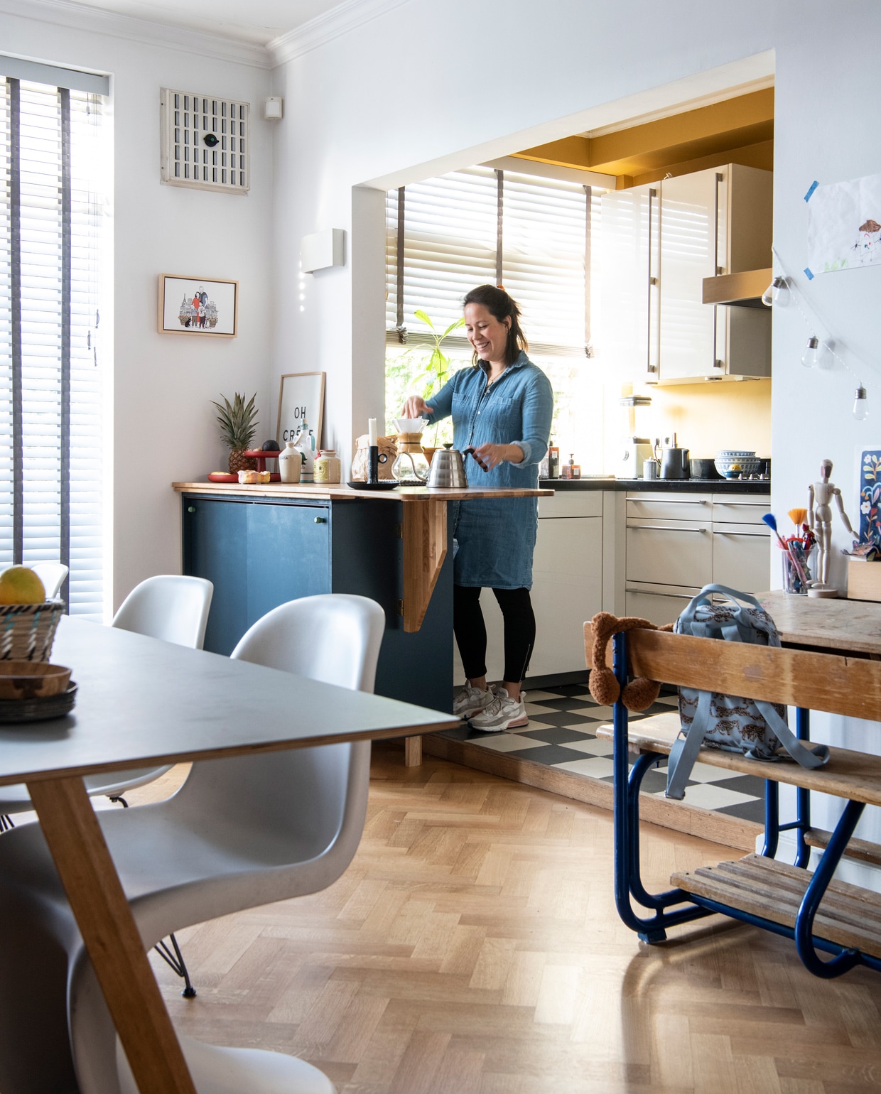 Keuken Siematic, muurverf India Yellow van Farrow &amp; Ball via De Ru. Vloertegels van winkel die niet meer bestaat. Kast op maat laten maken door een timmerman, zelf geverfd in kleur RAL 5008. Kastknoppen Oyoy. Wandlamp zat al in het huis. Oh Crêpe-poster Sissy-Boy, illustratie Clémentine Latron. Hoge pot souvenir uit Italië, mosterdpot Vendejo. Koffiepot Chemex, kandelaar souvenir uit België, waterkan Hario, fruitplateau Vitra, schaaltje knutselwerk Magalie, theepot Bredemeijer, schalen op aanrecht van Chinese toko. Tafel CPH30 van Hay, borden op tafel Serax, rieten mand souvenir uit Sardinië. Panton-stoel en Eames DSR-stoel Vitra. Vintage schoolbureau Marktplaats, tas Artis, potloden Van Gogh Museum. Ledlichtslinger Edison van Kikkerland. Overig onbekend.