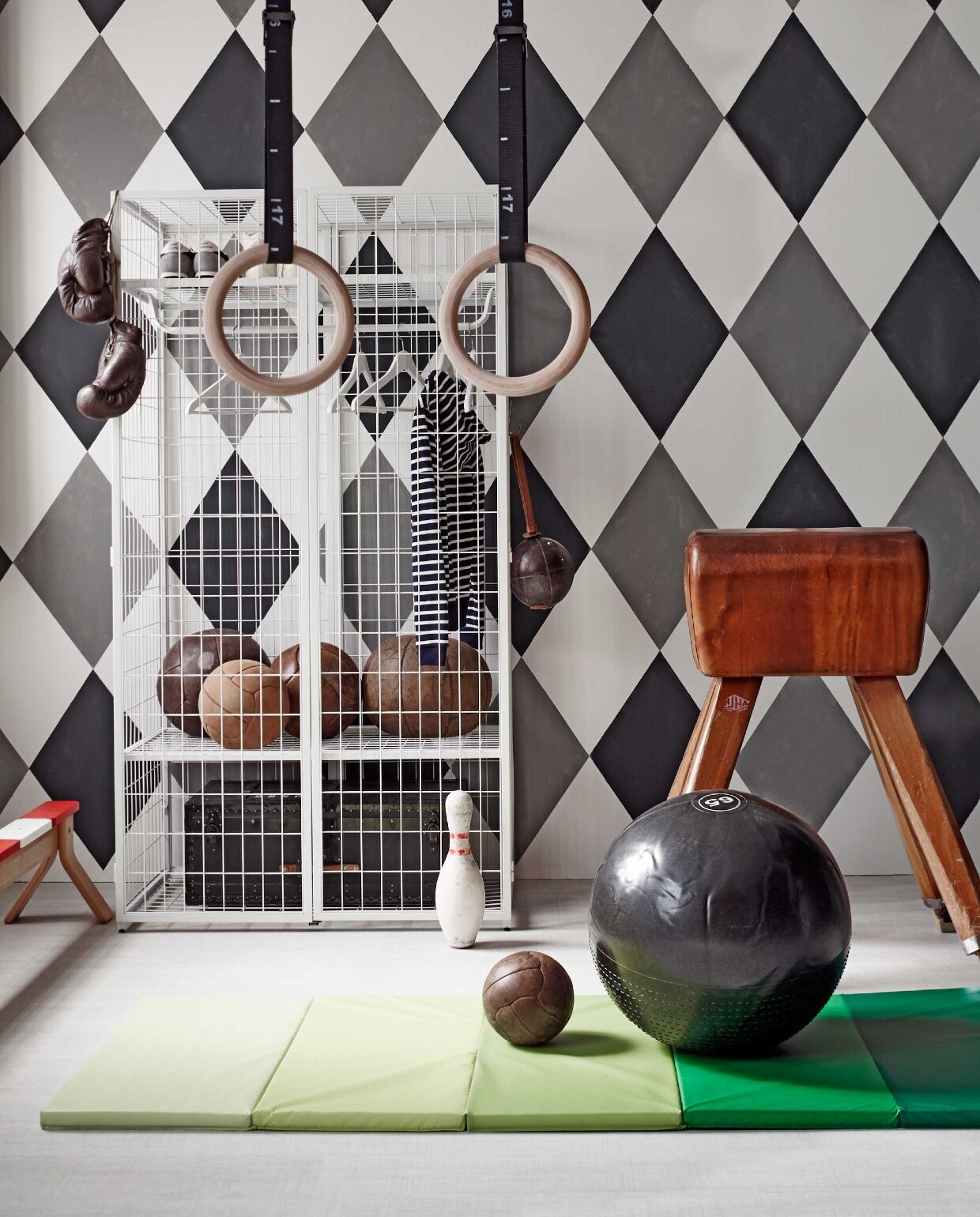 Kast Ikea PS 2014, gymmat Plufsig, evenwichtsbank Ikea PS 2014, set kledinghangers Bumerang (Ikea). Oude gymbok (Brût Amsterdam). Houten gymringen MP122 en gymbal van Adidas Ø 65 cm (www.muscle-power.nl). Oude leren gymballen en leren bokshandschoen (Spoor 38). Leren trainingsbal met leren lus en kegel (Neef Louis). Zwarte kist in kast (Combitex). Het dessin is geschilderd in muurverf grijs Plummett 272, zwart Off-Black 57, wit Strong White 2001 (Farrow &amp; Ball). Gestreept vest (Petit Bateau). Gympen New Balance (Maison NL).