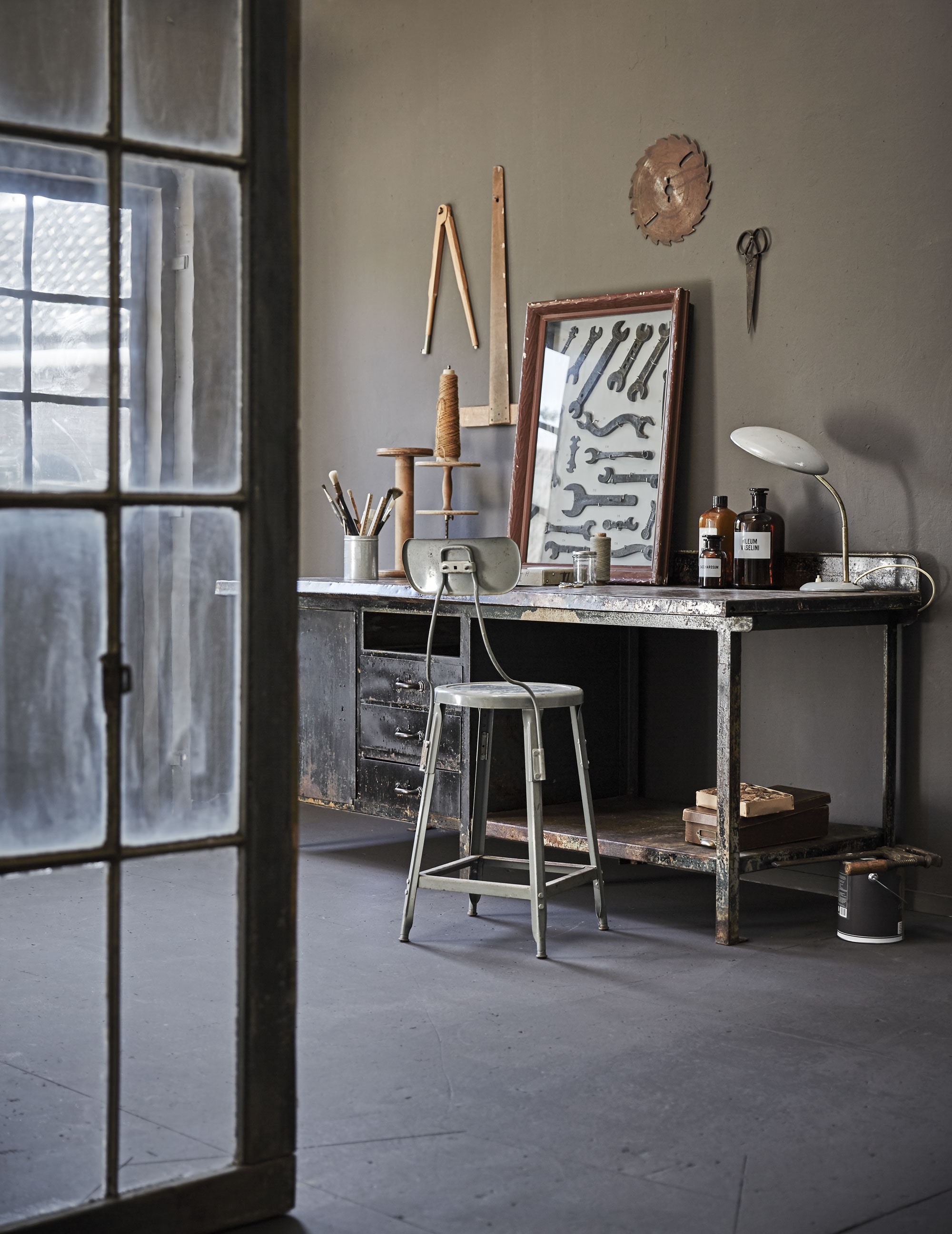 Fabrieksdeur met roedeverdeling (Combitex). Werktafel Stochov 75x197x80 cm (Brût Amsterdam). Werkplaatskruk (Spoor 38). Op het bureau: blik met kwasten, houten klos, garenhouder, ingelijste sleutels, bureaulamp (Spoor 38), apothekersflessen, schaar (Brût Amsterdam), doosje met garen (Combitex). Onder het bureau: bruine doos (Spoor 38). Aan de muur: passer (Six and Sons), liniaal (Neef Louis). Het overige is privébezit. Muurverf Mud Grey van vtwonen (vtwonen.be).