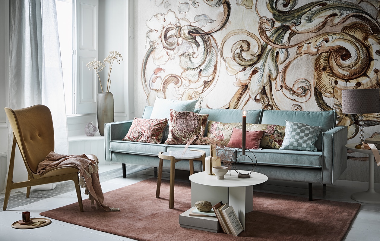 Behang Baroque R1151 (rebelwalls.com). Zeekleurige fluwelen driezitter Rodeo van BePureHome, 277 cm (vtwonen.be). Deense fauteuil links, wit met gouden vaas op tafel, lampvoet rechts naast de bank, kaars, ronde kandelaar op tafel, mok op de grond, plaid op de fauteuil (loods5). Witte stof Ditte voor het raam (ikea). Kussens van links naar rechts alle stoffen per meter: Manipur van Designers Guild groot kussen van Designers Guild, kussen met bedrukte stof van Nobilis, langwerpig kussen van Nobilis-stof, blauw kussen, stof van Osborne &amp; Little, klein roze kussen Rivoli van Designers Guild (wva). Lampenkap van stof Brooks van Zoffany, (barbaraemmy). Tafel met marmeren blad (bolia.com). Lage witte salontafel, roze kleed 140×200 cm, roze en blauw notitieboek, voorraaddoos op de grond, sierobject in raamkozijn (hay-amsterdam). Kom op salontafel (zarahome). Matglazen vaas met takken (homestede). Overig privébezit.