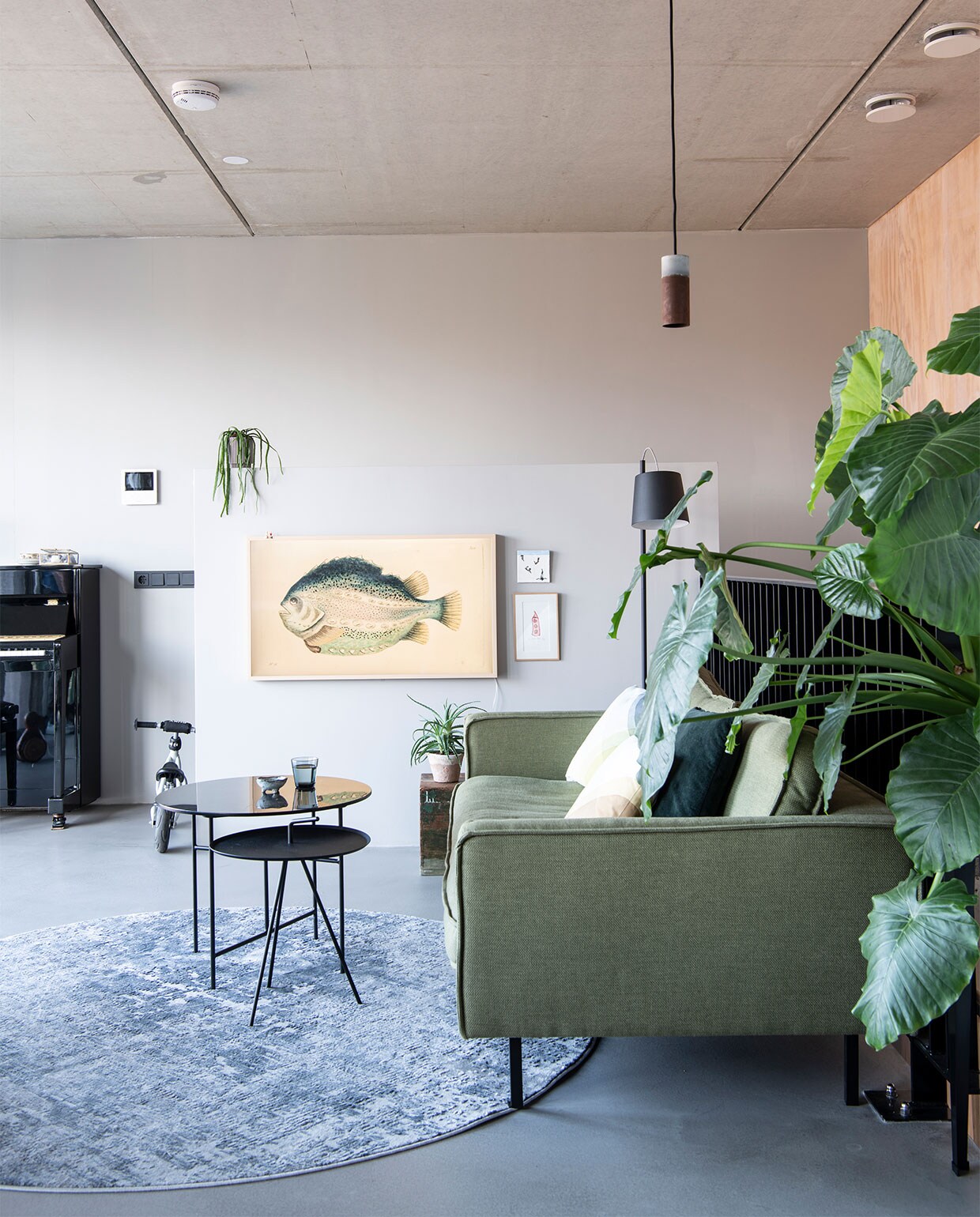 Pandomo K3 stuc als variant op een gietvloer. Bank Loods 5 Design. Herringbone kussens van Vitra via Groen + Akker. Groen kussen en plantenstandaard Intratuin. Vloerkleed Gusta. Hoge bijzettafel Loods 5. Lage bijzettafel Woood. Vloerlamp Zuiver. Hanglamp Roest van Van Joost voor Graypants. Tv Samsung Frame met camouflagebeeld vis. Tegel StoryTiles. Schilderij gekocht in een Parijse galerie.