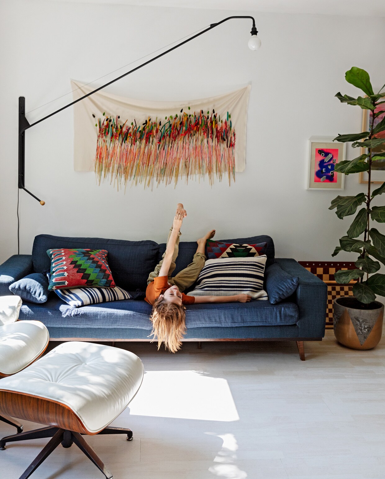 Bank Wehkamp. Kussens Ikea en Arket. Wandlamp Potence van Vitra. De witte lounge chair met Ottoman is een kopie van Eames. Wanddoek met geverfde grassen eigen werk. Pot goud/grijs Van Pot Los. Zeefdruk Tim Wilders/Atelier Haven. Schaakbord meegenomen uit Londen.