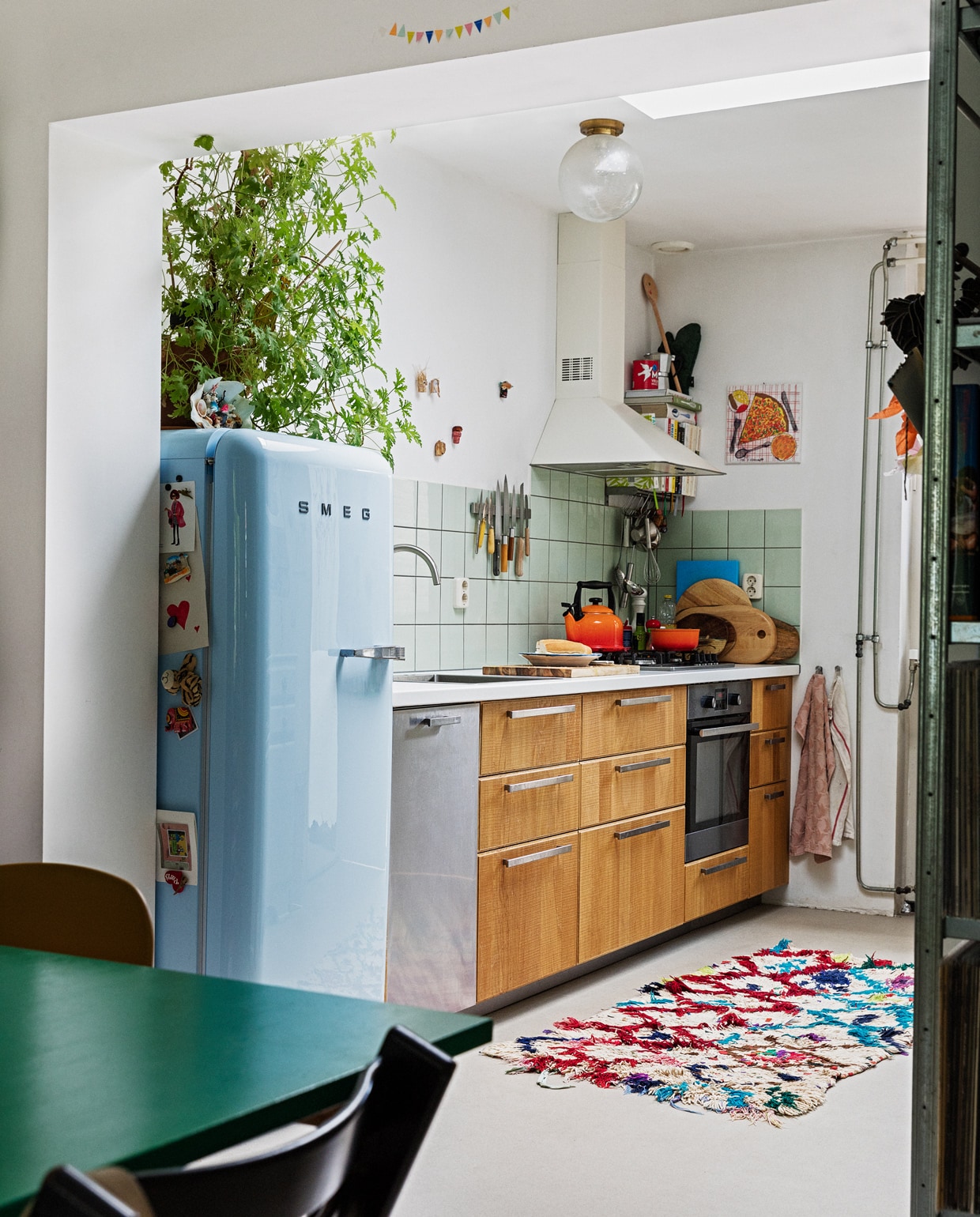 Koelkast Smeg. Geraniumplant zelf gestekt. Kitsch schelp op koelkast souvenir uit Griekenland. Magneet-souvenirs meegenomen van verschillende reizen. Fluitketel en pan Le Creuset. Messen Suncraft, Opinel, Dille &amp; Kamille, Pallarès Solsona. Snijplanken Hay, Ikea, Serax. Soeplepel Brabantia. Rvs keukengerei Dille &amp; Kamille. Schaaltje van een pottenbakkersmarkt. Afzuigkap en oven Ikea. Ovenhandschoen Kook. Pollepel Kookhuis aan de Maes. Handdoeken Sissy-Boy. Tapijt The Souks. Pizza-schilderij Oskar. Ronde plafondlamp lampenwinkel.nl.