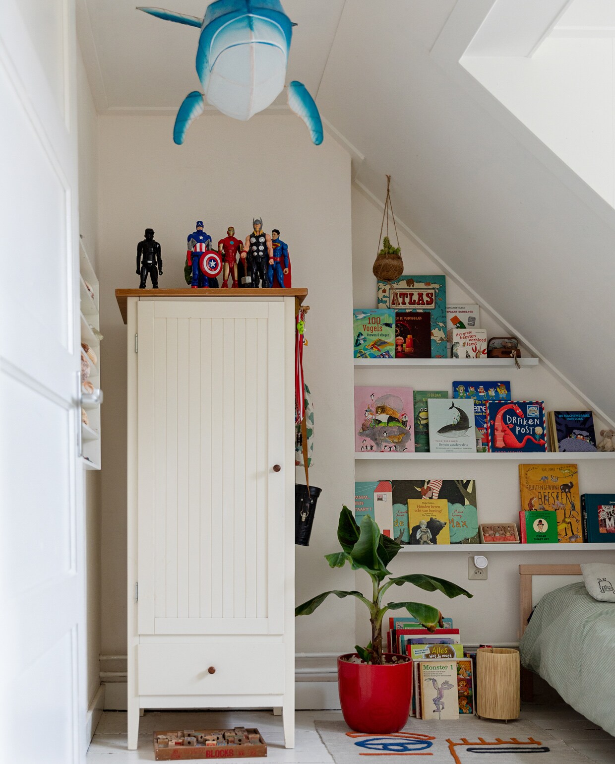 Papieren walvislamp Petit Pan. Witte kast, rode pot, houten letterdoos, letterbak aan de wand en Marokkaanse lamp op de grond kringloop. Tapijt Kwantum. Verzameling superhelden van de rommelmarkt. Bananenplant Intratuin. Bed en beddengoed Ikea. Grijs kussen met vogels Zilalila. Vintage verrekijker van opa.