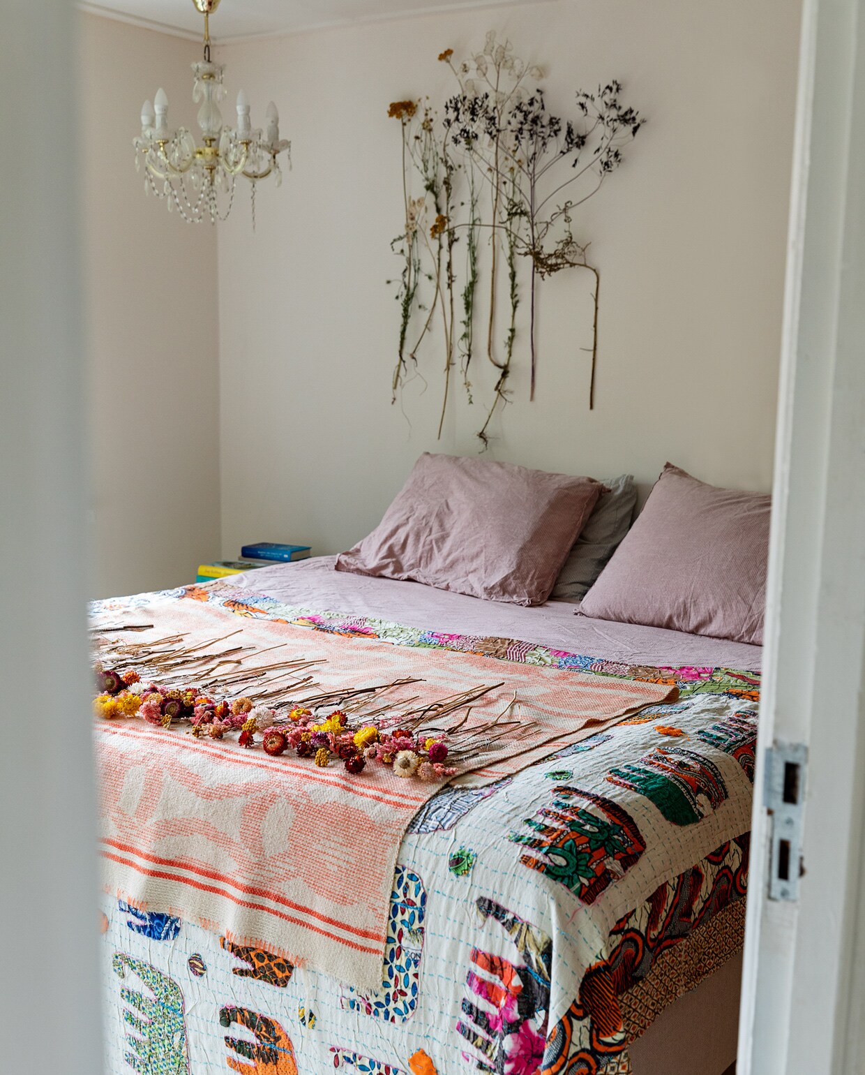 Bed en beddengoed Ikea. Deken met olifanten souvenir uit India. Kroonluchter Sissy-Boy. Roze plaid met gedroogde bloemen en verzameling gedroogde planten aan muur Loesje van Herp.