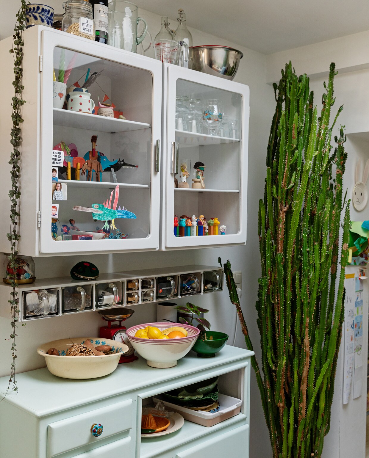 Vitrinekasten en kruidenrek Piet Zwart. Dressoir kringloop. Schalen en kommen Tine K Home en In-Petto. Weegschaal Dille &amp; Kamille. Mengkom rvs Duikelman. Grote cactus Marktplaats. Hangplant Ceropegia Intratuin. Pot kringloop. In de kast: verzameling Pez-snoephouders. Kartonnen dieren zelfgemaakt. Wijnglazen Iittala. Aardewerk meegenomen van diverse reizen. Tekeningen en rariteitenkabinet van Oskar.