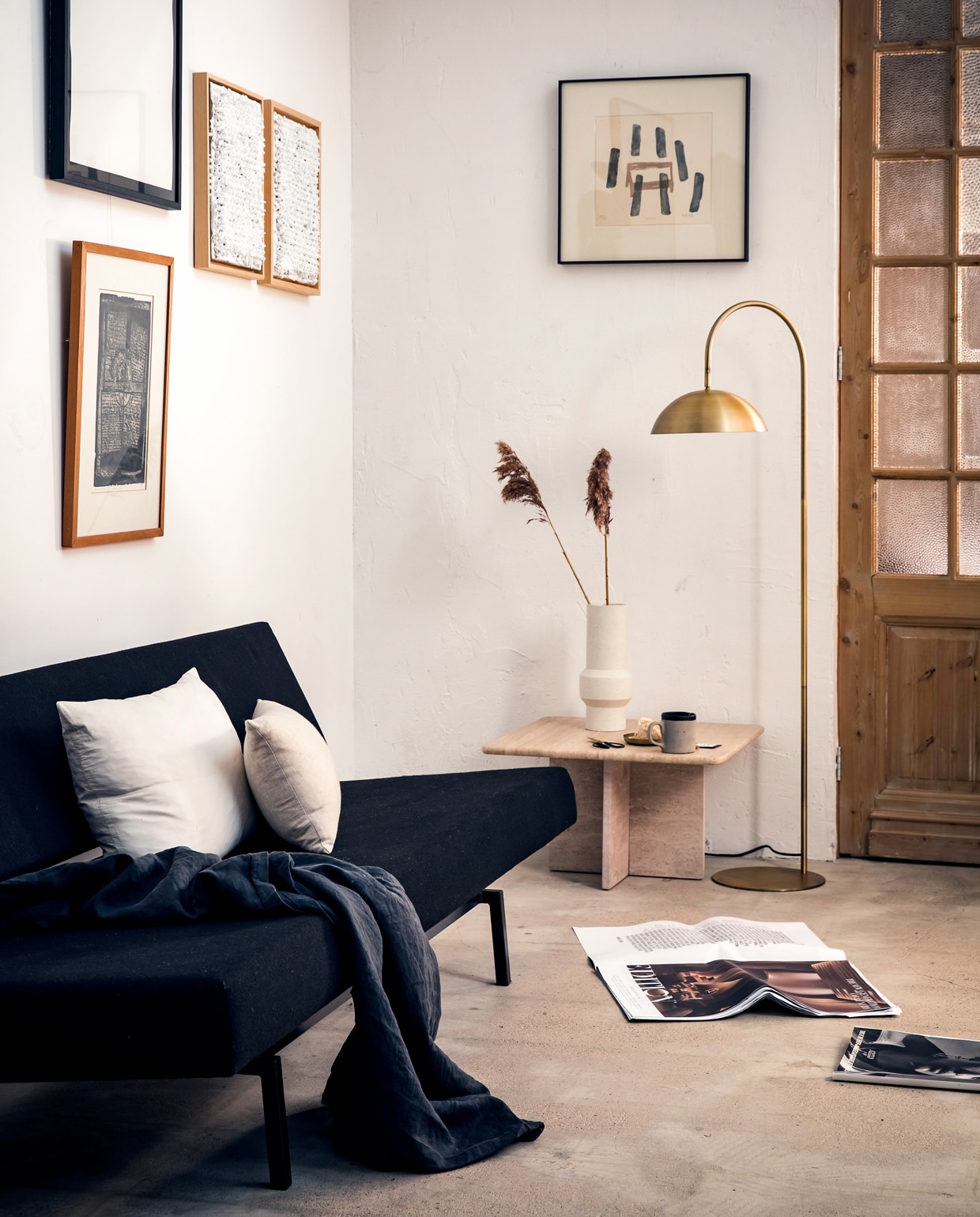 Vloerlamp Jupiter* brons met antiekfinish, 144×47,5 cm (hxb), kap Ø 28 cm, voetplaat Ø 28 cm. Ook in deze serie, allemaal in brons met antiekfinish: tafellamp Jupiter*, 51×28 cm (hxb), kap Ø 20 cm, voetplaat Ø 18 cm, klemlamp Jupiter*, 78×30 cm (hxb), kap Ø 20 cm, wandlamp Jupiter*, 68×50 cm (hxb), kap Ø 20 cm. *met led-verlichting. M.m.v. cocooncollectables.nl (vintage slaapbank, tafel van travertin, lijsten), hkliving.nl (vaas), hm.com (zwart laken, wit groot kussen), bymolle.com (langwerpig linnen kussen), overig privébezit.