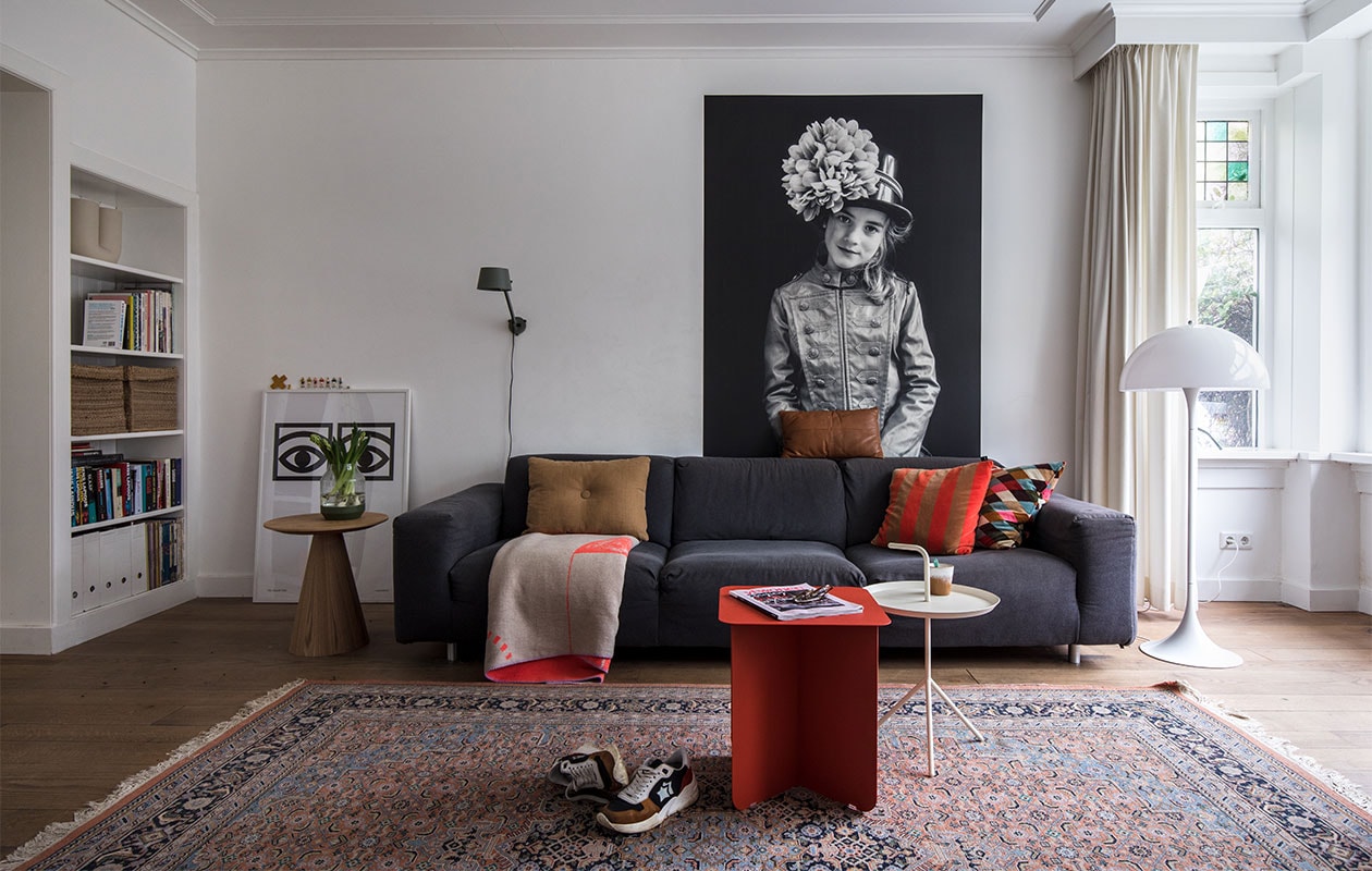Print links: Ögon Cacao van de Zweedse grafisch ontwerper Olle Eksell. Bijzettafel Kwantum. Vaas Muuto. Wandlamp Tonone Bolt. Bank gekocht tijdens een garagesale. Plaid op bank Adelheid Laden. Kussens: bruin Hay, cognac Leen Bakker, gestreept Hay, wybers Ikea. Print achter bank foto van dochter Meis gemaakt door Maaike en geprint op Gekkotex, een zelfklevend polyester. Vloerkleed kringloop. Sneakers Atlantic Stars. Rode bijzettafel Puik Hinge. Witte bijzettafel Hay. Mok HKliving. Vloer van Amerikaans eiken lag er al. Muurverf wit RAL 9016 van Sikkens.