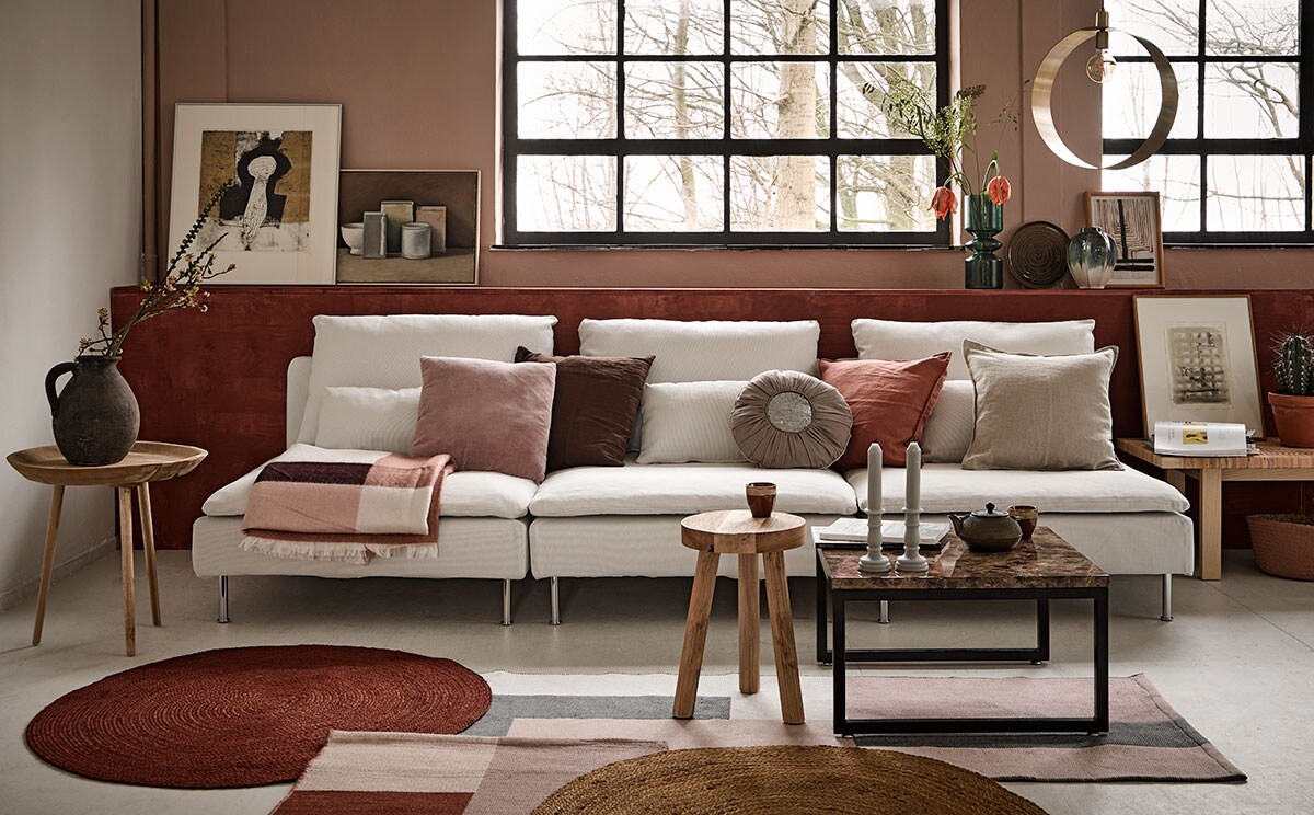 Wand geschilderd in Skin Powder, lambrisering in Cardinal Red, matte krijtverf (Pure &amp; Original). Bank Soderhamn, tafel Stockholm (Ikea). Op de bank kussens v.l.n.r. roze (Trendhopper), bruin (Ikea), rond (Loods 5), gewassen linnen in de kleur roze en écru, plaid, wollen vloerkleden met blokpatroon (H&amp;M Home). Ronde juten vloerkleden, 3-poot kruk, bijzettafel van gerecycled hout (Kwantum). Salontafel marmerlook, goudkleurige metalen hanglamp Yane, op lambrisering hoge glazen vaas Bali, tweekleurige vaas, mand onder sidetable (Trendhopper). Decoratiebord (Kwantum). Cementen vintage kruik, goudkleurige Japanse theepot, kommetjes, kaarsen (Loods 5). Alle kunst komt van de SBK Kunstuitleen Amsterdam.