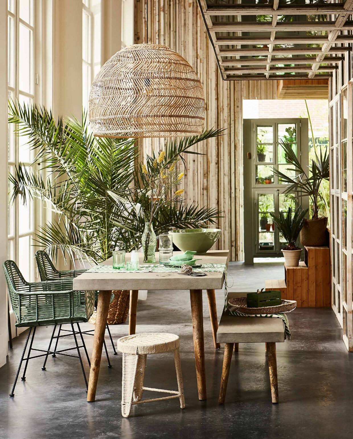 Tafel met betonnen blad en bijpassende bank (Nordal). Gevlochten hanglamp, groene rieten stoel (HK Living). Kruk van gevlochten touw, gevlochten schaal (Madam Stoltz). Lopers, groen met wit en groen kratje (H&amp;M Home). Karaf in de vorm van een vis, groene visvormige glazen schalen, lage visschaal van glas, laag groen glas, hoog groen glas, wit zoutvaatje, alles uit de collectie van Paola Navone (Serax). Glazen karaf met ribbel (Household Hardware). Grote groene aardewerken schaal (De Weldaad). Rieten mand met palm, gevlochten mand met palm op trap (Nordal). Groene kast (Combitex). Overig privébezit.