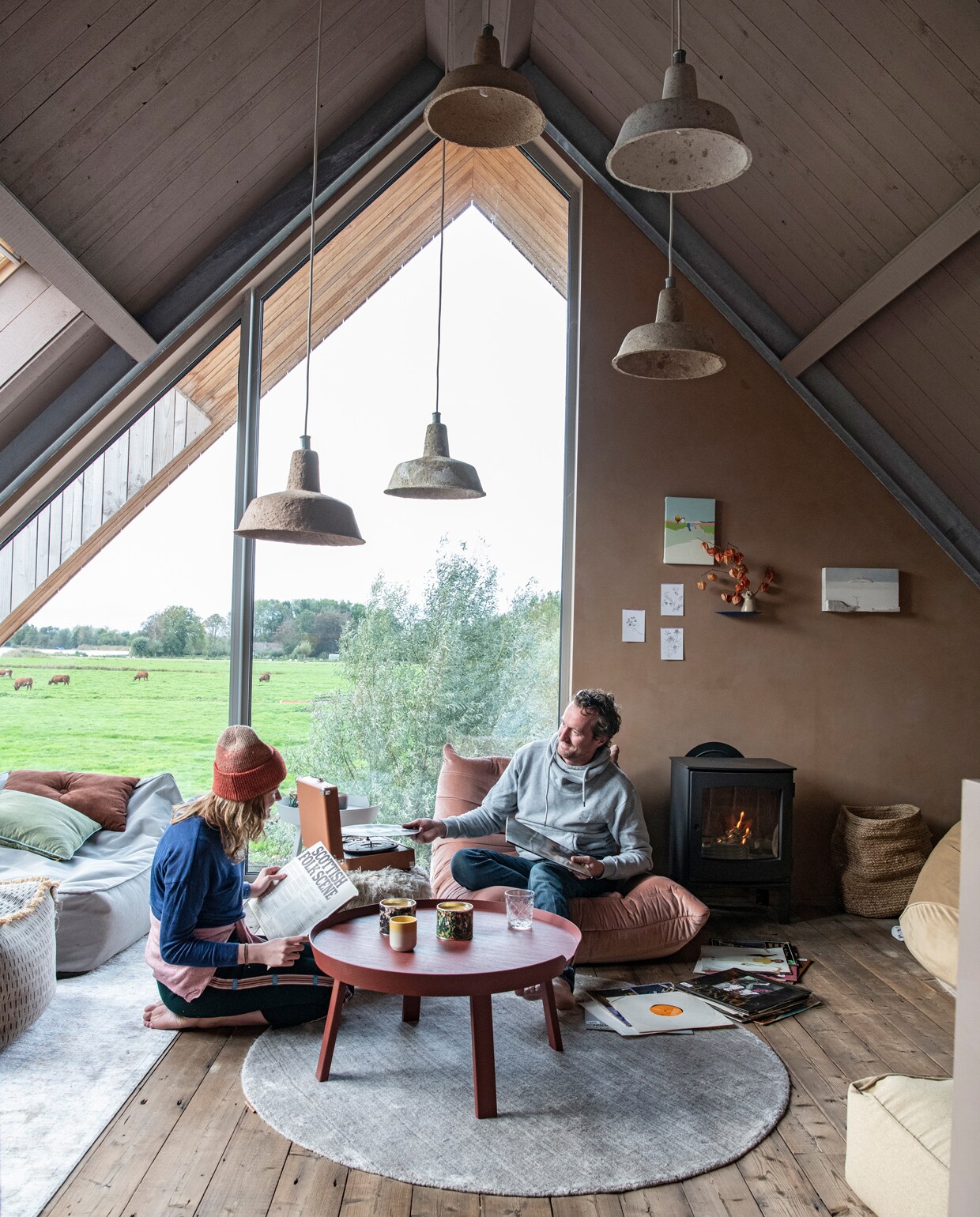 Blauwe en beige zitzakken en harige poef Vetsak. Kussens Wehkamp. Roze stoel Bestwelhip. Salontafel Muuto. Glas Ikea. Kaarsenhouder Sissy-Boy. Hanglampen van Ania Werner. Houtkachel Dru. Mand Fonq. Wandplank Studio de Winkel. Schilderijen met huisjes van Donna Walker via Etsy. Kleden RugVista. Op de muur kalkverf in de kleur Topaze van Pure &amp; Original.